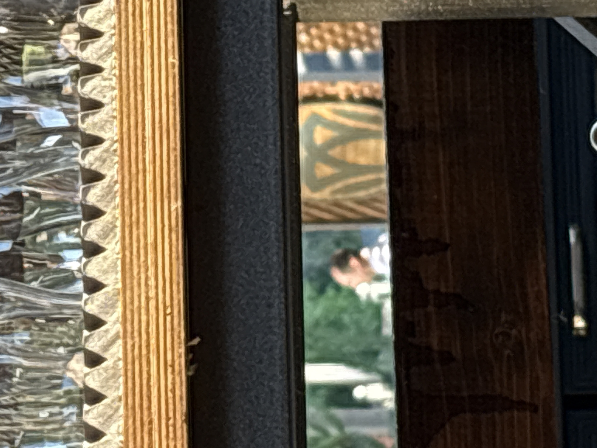 The image presents a close-up, vertically oriented composition, characterized by several distinct vertical sections.
On the far left, a textured surface with a ridged, possibly reflective pattern is visible. Adjacent to it is a light brown, corrugated material, likely a decorative element or part of a frame. Moving right, a dark, subtly patterned surface, possibly fabric or painted wood, occupies a significant portion.
Central to the image is a narrow, vertical reflective strip, appearing to be a mirror or a window pane. This strip captures a blurred reflection: the upper part shows an intricate, patterned surface, possibly an ornate ceiling or building exterior, with shades of orange, brown, blue, and green. Below this, green foliage is reflected, indicating an outdoor setting. The lower section of the reflection clearly shows the head and shoulders of a person, wearing a light-colored top, bending forward or looking downwards amidst the green background.
To the right of this reflective section, a dark wooden surface with a visible grain pattern fills the space. The far right edge of the image reveals a slim, dark vertical strip, with a small, bright, indistinct object near the top right corner and a longer, thin bright element positioned lower down, possibly a piece of hardware or a fixture.
The scene depicts a daytime setting due to the bright lighting. The primary activity is the reflection of a person seemingly engaged in an outdoor activity, possibly gardening or inspecting something low to the ground. The combined elements suggest a view or reflection originating from an indoor space looking out onto an outdoor environment. No text is visible in the image.