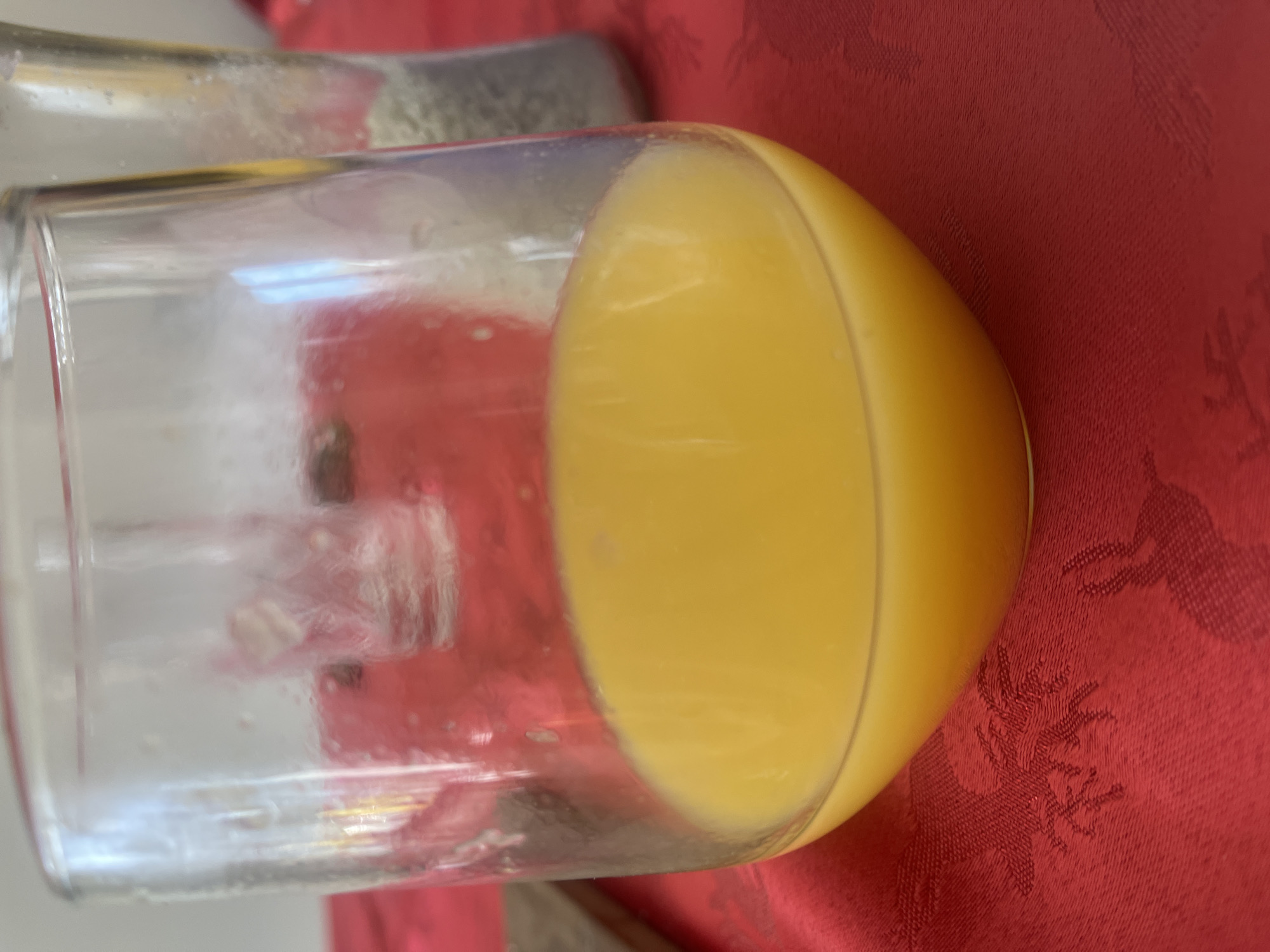 A glass containing yellow liquid, possibly orange juice, is resting on a red tablecloth. The tablecloth has a woven pattern. The glass is clear with visible condensation. The scene is likely indoors. The location cannot be determined based on the visual data. No people, subjects, objects, actions, or interactions beyond the presence of the glass and tablecloth can be ascertained.
