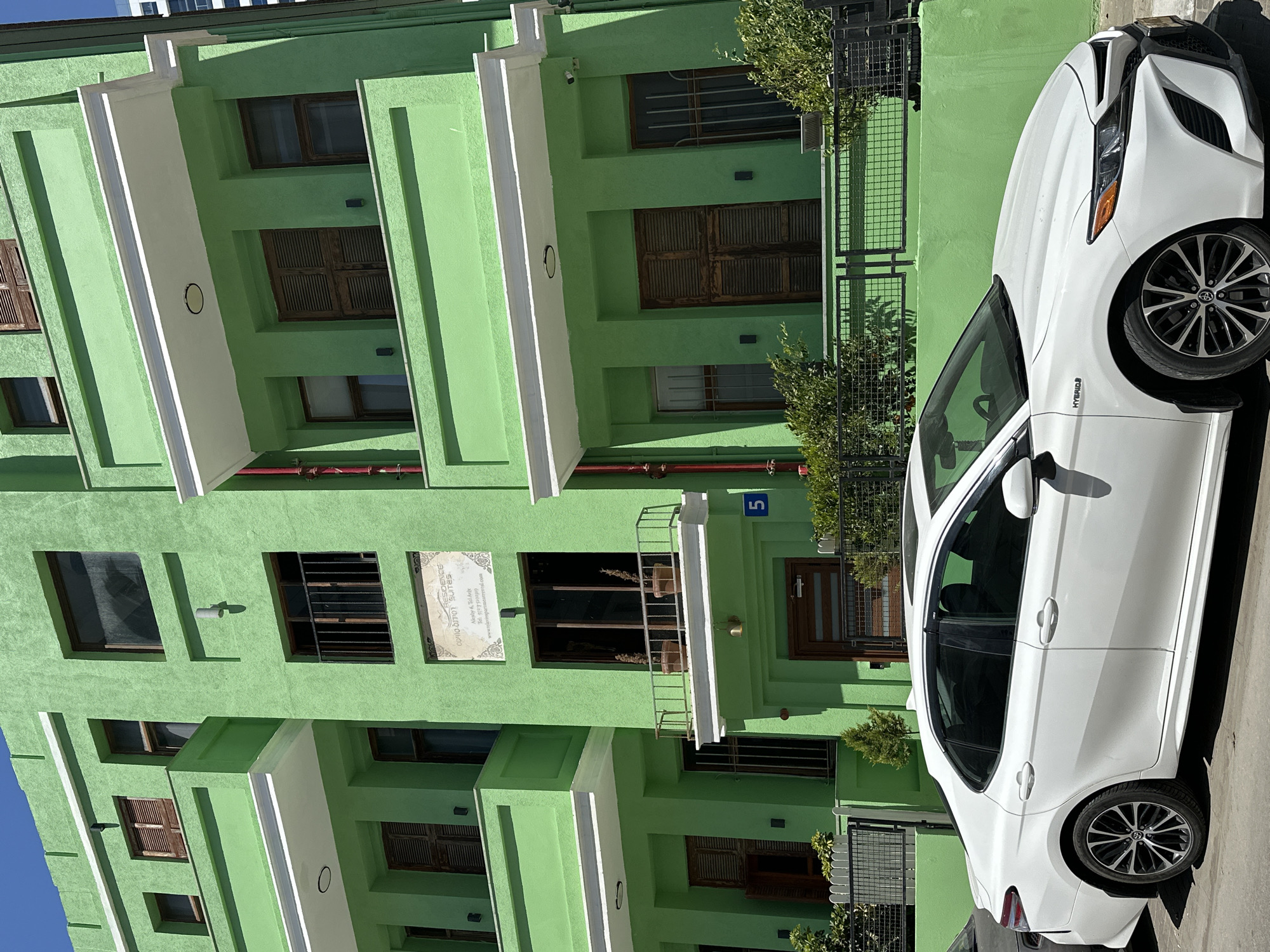 An eye-level shot captures a vibrant green multi-story building alongside a white car, likely on a street in Tel-Aviv, Israel. The building, painted in a distinctive bright lime green, features several rectangular windows, some with dark wooden shutters or metal grates. White rectangular ledges or balconies extend from the facade on various levels, creating a tiered effect.
A white Toyota Camry Hybrid is parked on the street in front of the building, occupying the right side of the frame. The car faces left, showcasing its front end and side profile, with "HYBRID" visible on the fender. Its wheels feature a black and silver spoke design.
Notable details on the building include a small blue sign with the number '5' displayed near a dark wooden entrance door. Higher up, a white rectangular plaque with black text reads "DOUZOI RESIDENCE SUITES", with additional lines below including "Ahuzat Tel Aviv", a partial phone number, and a website "www.telavivresidencesuites.com", indicating the building's function as a suite or apartment complex. A black metal fence or gate with lush green bushes runs along the ground level, separating the building from the street.
The scene is brightly lit by natural sunlight, casting distinct shadows and suggesting a clear, sunny daytime. No people or overt activities are visible, presenting a static urban landscape.