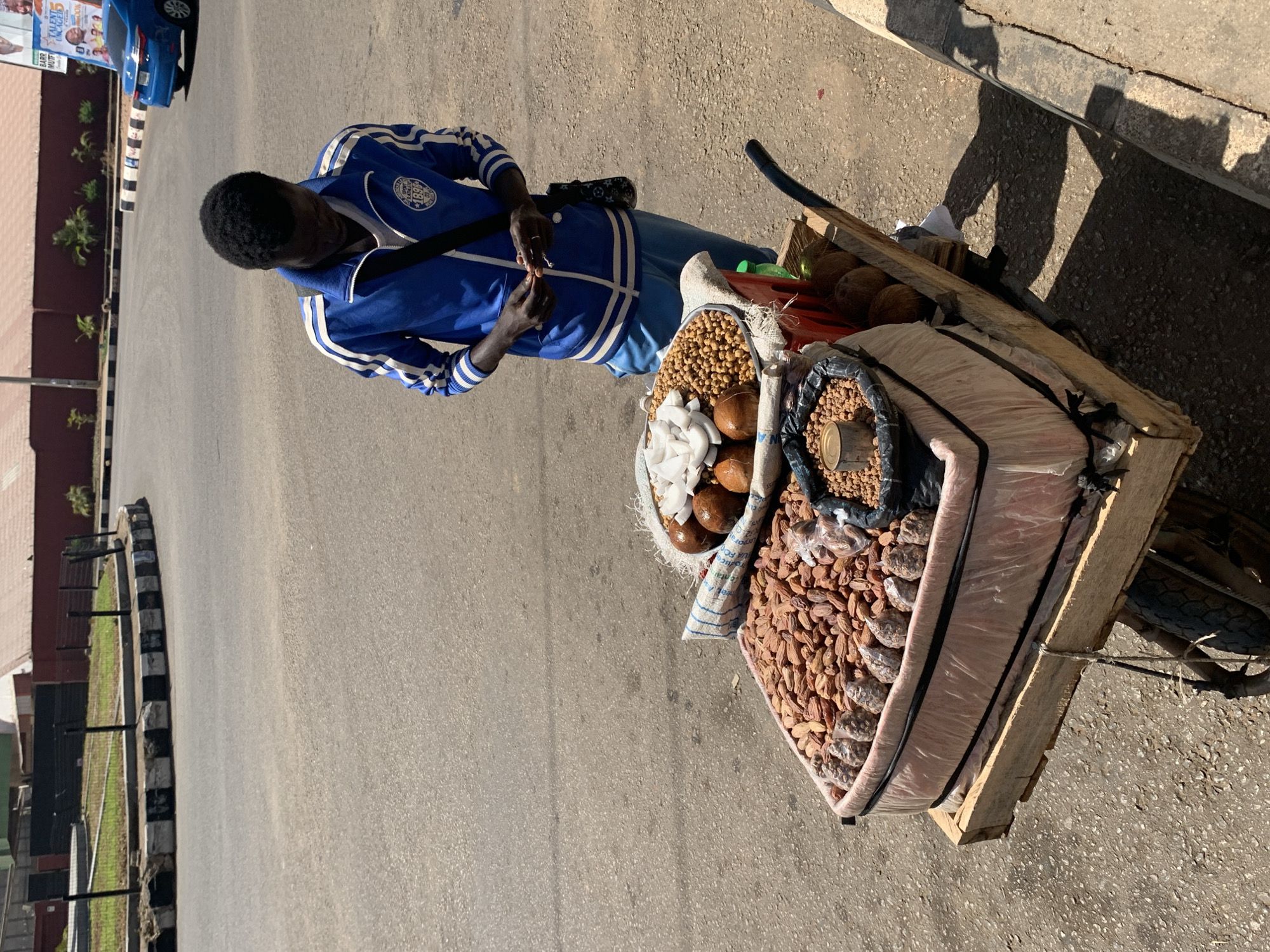 An overhead shot depicts a vendor and a bicycle laden with goods, likely in the city of Jos, Nigeria. The vendor, a dark-skinned male, is wearing a blue tracksuit and stands on a paved road. He appears to be focused on something in his hands, possibly a phone or money. The bicycle has a wooden crate attached to the back, which is filled with various food items including coconuts, peanuts, and other nuts. The road has a curved section to the left with a black and white painted curb and a green grassy area behind a black fence. To the right, the road meets a curb. The sun casts shadows from the bicycle and the adjacent curb. A building is visible in the background on the left. A blue vehicle is also visible in the upper left.
