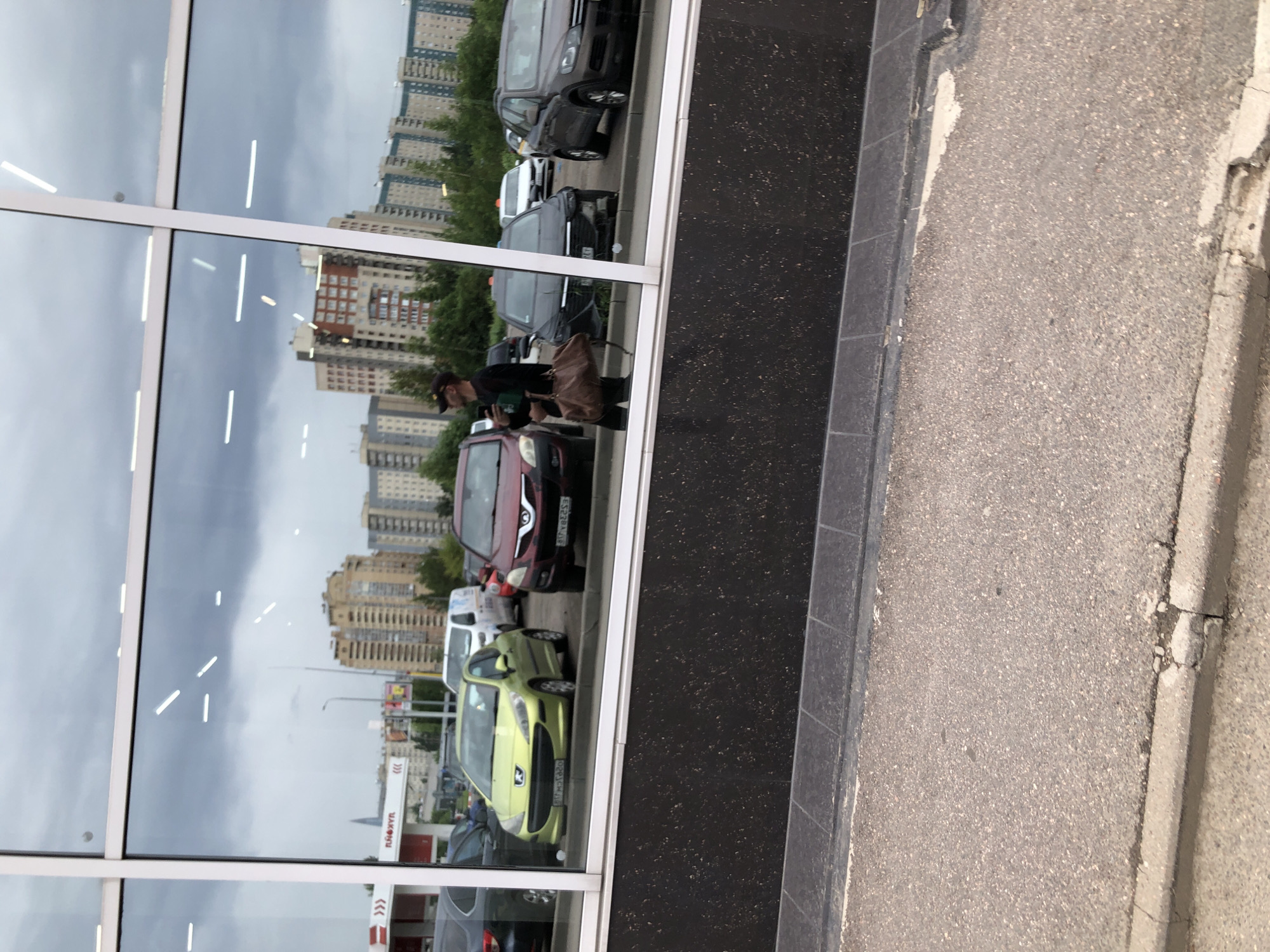 The image shows a street scene reflected in a large window. In the reflection, a person is walking with a brown bag and a phone. Several cars are parked or driving, including a green Peugeot 207 with license plate BC3322OA, a red car, and a dark gray car. In the background, there are apartment buildings, trees, and a gas station with the logo "Lukoil." The sky is overcast. The asphalt road and a curb are visible in the foreground. Based on the "Lukoil" gas station and the architecture, this scene is likely in Saint Petersburg, Russia.