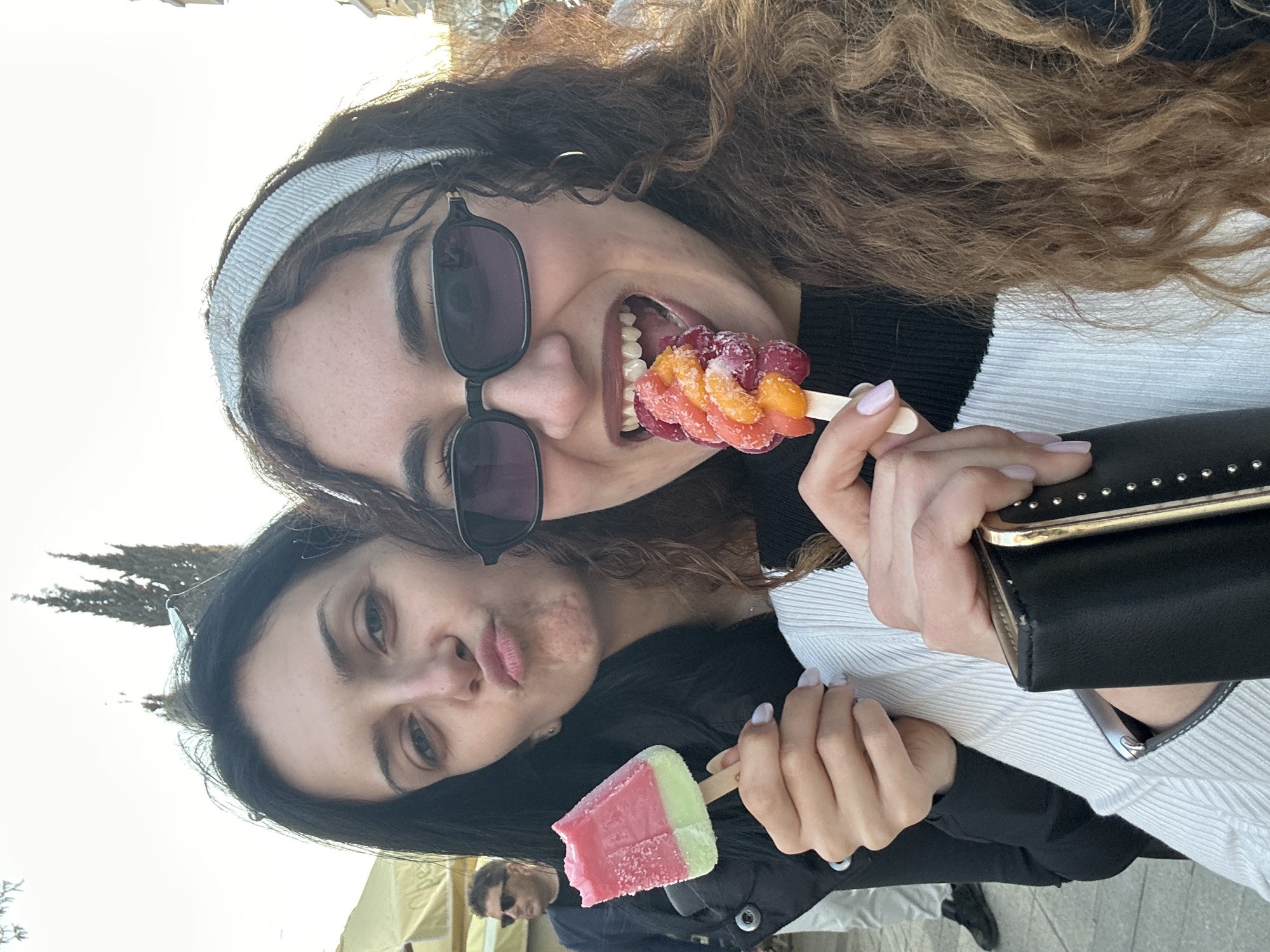 Two women pose with frozen treats. The woman at the top has curly hair, sunglasses, and a gray headband, and she is holding a fruit skewer in her mouth. The woman below has dark hair and is puckering her lips. She holds a pink and green popsicle. The woman at the top wears a black and white ribbed shirt and holds a black purse. The woman below wears a black jacket. A partially visible man in sunglasses is in the bottom left corner. The location is the city of Burgas, Bulgaria.