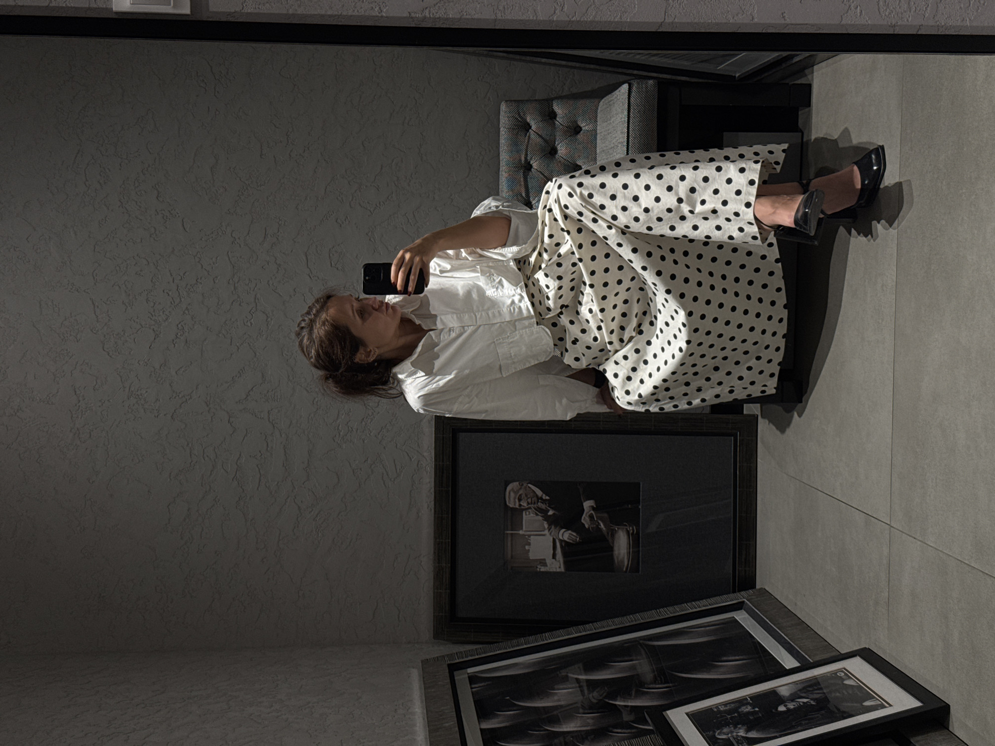 This image captures a woman seated indoors, engrossed in her smartphone. She is positioned on a grey, upholstered armchair or small sofa, with her body angled towards the left. Her dark hair is pulled back, and she holds a black smartphone in her right hand, appearing to take a selfie or interact with an application.
She wears a white long-sleeved shirt and a flowing white skirt or trousers adorned with black polka dots. Her feet, encased in shiny black high-heeled shoes, are visible to the right of the chair.
The setting features a textured grey wall and a floor composed of large, light grey tiles. Several framed artworks are arranged on the floor in the foreground. One prominent frame displays a black and white image of an older man in a suit, while another features an abstract or geometric black and white pattern. A third, smaller framed picture is partially visible. The overall color palette is minimalistic, dominated by shades of grey, white, and black.
The lighting is soft and somewhat dim, casting subtle shadows that emphasize the textures of the wall and clothing. The woman's expression is neutral and focused on her device. The scene conveys a moment of personal interaction with technology within a stylish, contemporary interior. Although no explicit location cues are visible in the image, the context provided situates the scene in Dubai, United Arab Emirates. No visible text is legible in the image.