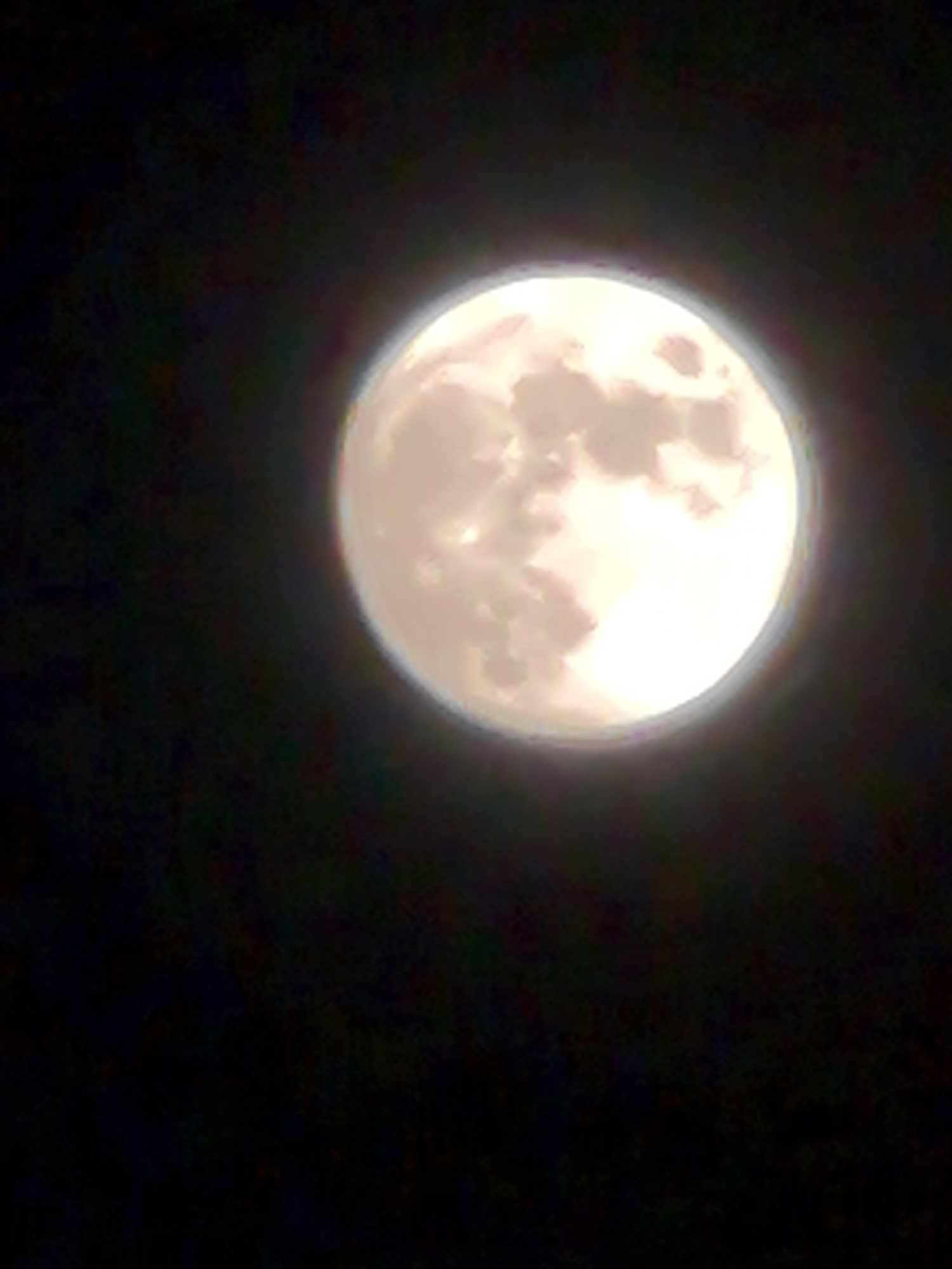 A full moon dominates the upper right quadrant of the image, set against a dark, presumably night sky. The moon's surface displays varying shades of light and shadow, indicating its craters and topographical features. There is a diffuse halo surrounding the moon, which might be attributed to atmospheric conditions or the camera's optics. The rest of the image is predominantly black, suggesting a dark, unobstructed view of the night sky. No specific details link this image to Noviy, Russia.