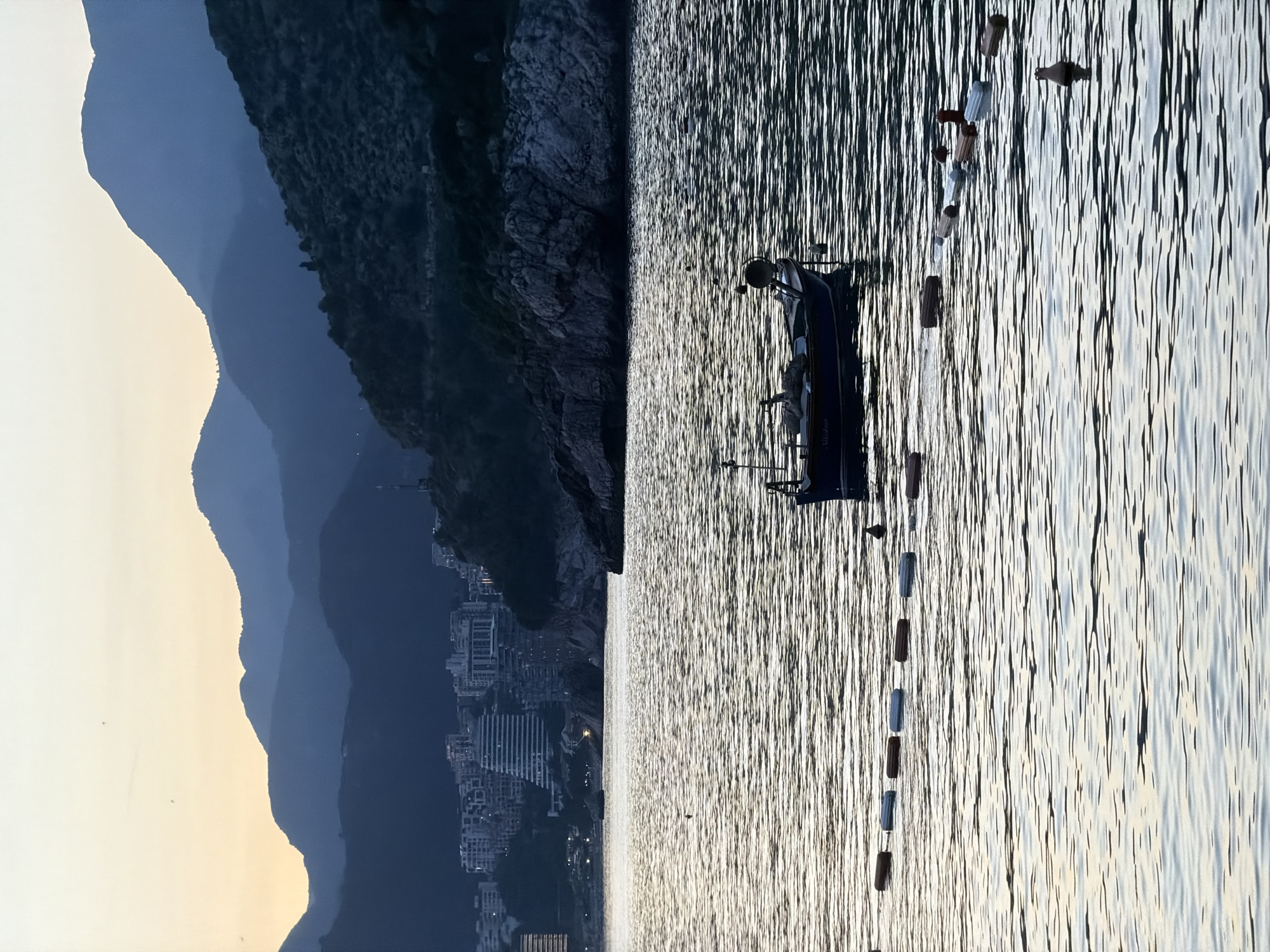 The image captures a dusk scene in Pržno, Montenegro. The landscape features a silhouette of mountains fading into the evening sky on the left. To the right, the sea reflects the faint light, displaying distinct wave patterns. A small boat is positioned in the water, attached to a string of buoys marking a boundary. A line of buildings is visible on the shore.