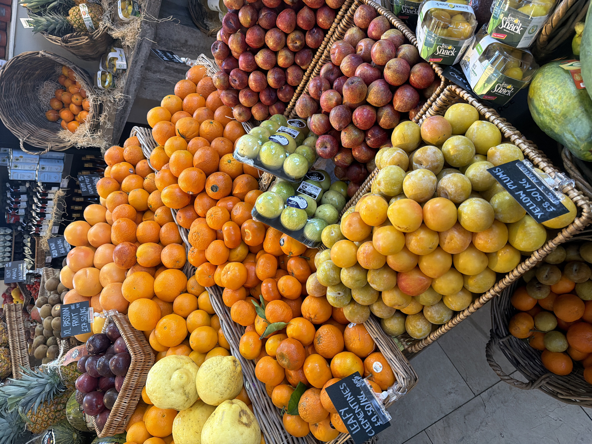 A vibrant display of fruits is captured in a close-up shot. Baskets overflow with oranges, plums in varying hues, apples, pineapples, kiwis, and passion fruit, all neatly arranged. Price tags are visible, indicating items like 'Leafy Clementines' at £9.50/KG, 'Kent Mango,' 'XL Passion Fruit,' and 'Yellow Plums' at £8.50/KG. The scene suggests a market or upscale grocery store, with the produce arranged for sale. The location is likely in London, United Kingdom, based on the currency (£).