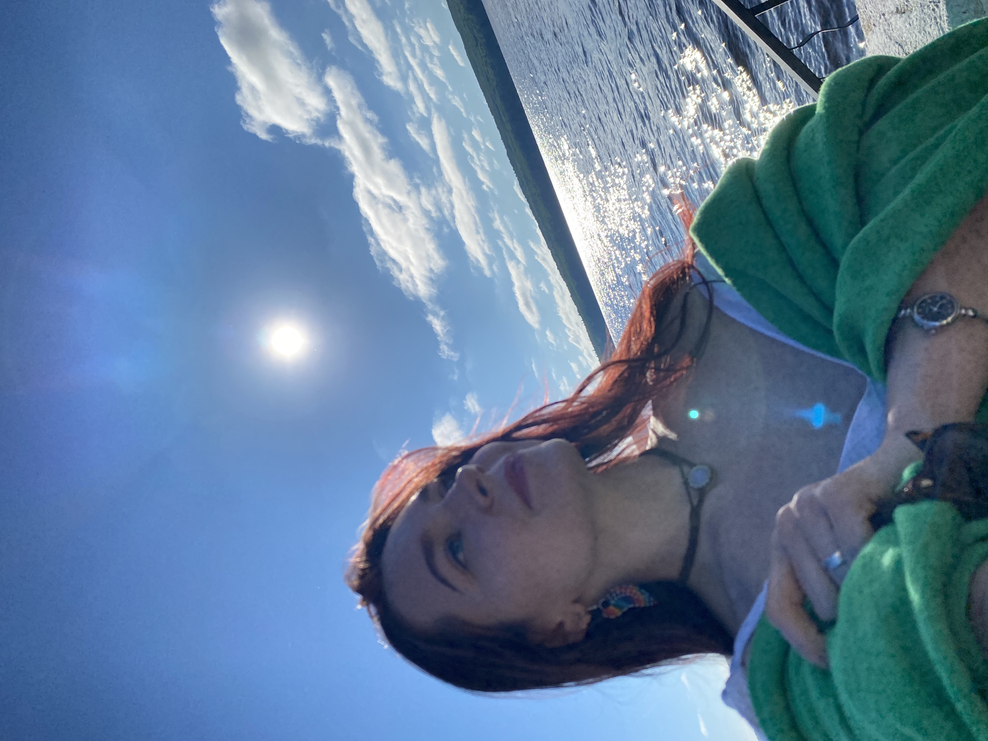 A medium shot captures a young woman with long, reddish-brown hair, wearing a green shawl, standing on what appears to be a pier or waterfront promenade. She is looking upwards and to her right, with a slight smile. The sun is bright and high in the sky, creating a lens flare. She is wearing a necklace with a pendant and a ring on her left hand. The background shows a body of water, likely a river or lake, with sparkling sunlight on its surface. A forested shoreline is visible in the distance, under a bright blue sky with scattered clouds. The location is identified as Chaikovsky, Russia.