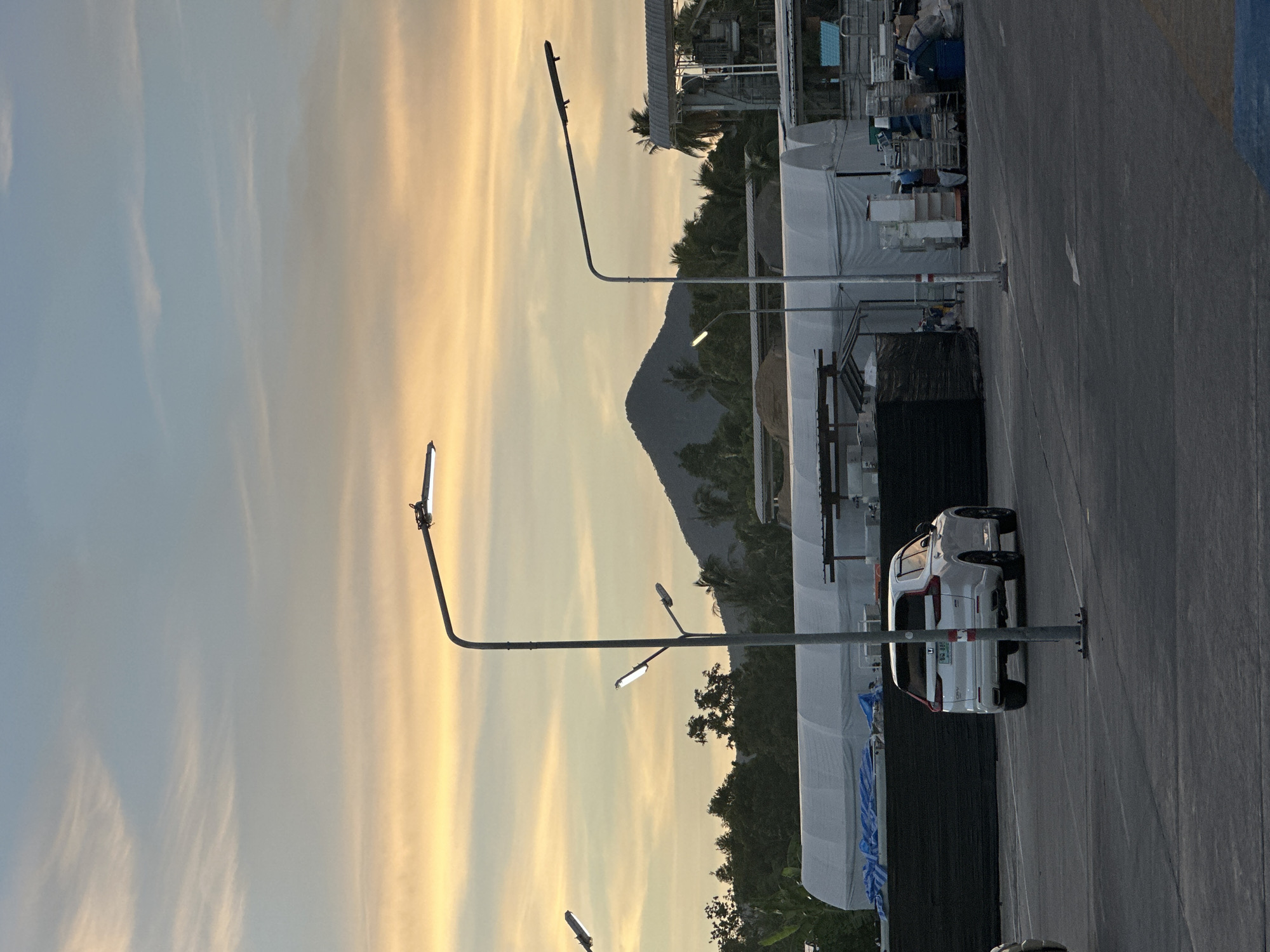 This is a vertical, eye-level medium shot of a parking lot at sunset. The sky is a mix of pale yellow and blue with wispy clouds. In the foreground, a white SUV is parked. In the background, there are some tents, palm trees, and a mountain. The scene is likely in Ko Pha Ngan, Thailand, given the location context. The lighting suggests it is late afternoon or early evening.
