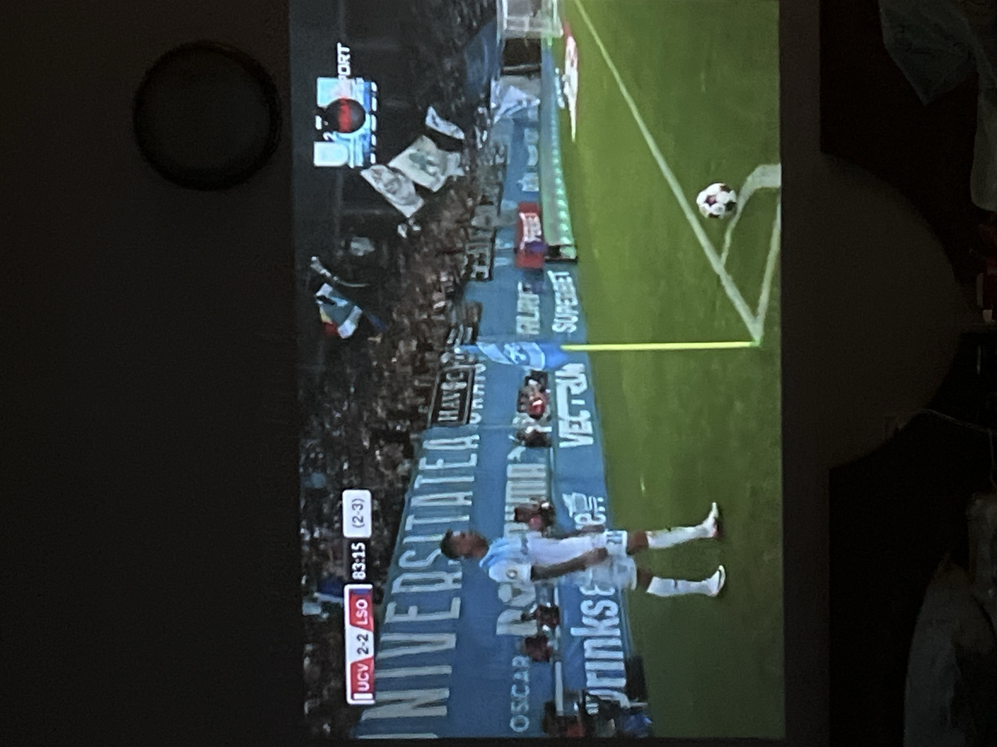 A projection of a soccer match is displayed on a wall. The score is 2-2, and the time is 83:15, with the aggregate score showing 2-3. A player in a light blue and white uniform stands on the field, facing away from the camera. The field is green, with white lines marking boundaries. A soccer ball is positioned near the corner flag. In the background, spectators fill the stands, and flags are visible. A banner with the word "UNIVERSITATIA" is also displayed. The projection is taking place in a dimly lit room, possibly a living room, as indicated by the furniture and dark surroundings. The specific location in Obzor, Bulgaria, cannot be determined from the image.