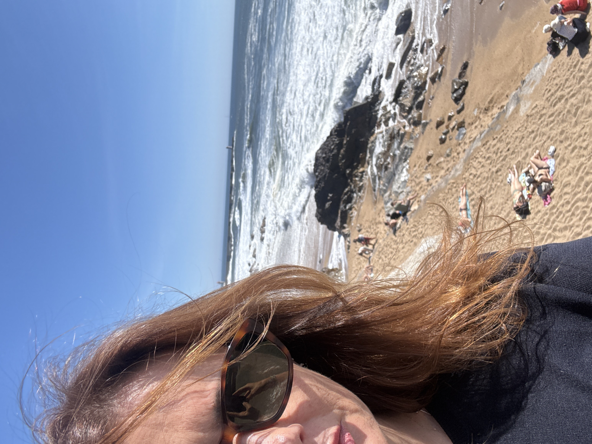 The image shows a view from a person lying on a beach. The beach is sandy with rocky outcrops and the ocean is visible in the background. There are other people on the beach in the distance, some sunbathing. The person in the foreground is wearing sunglasses and a dark-colored shirt. Based on the environment and the presence of a possible breakwater in the distance, it is possible that the location is one of the beaches near the city of Porto, Portugal.