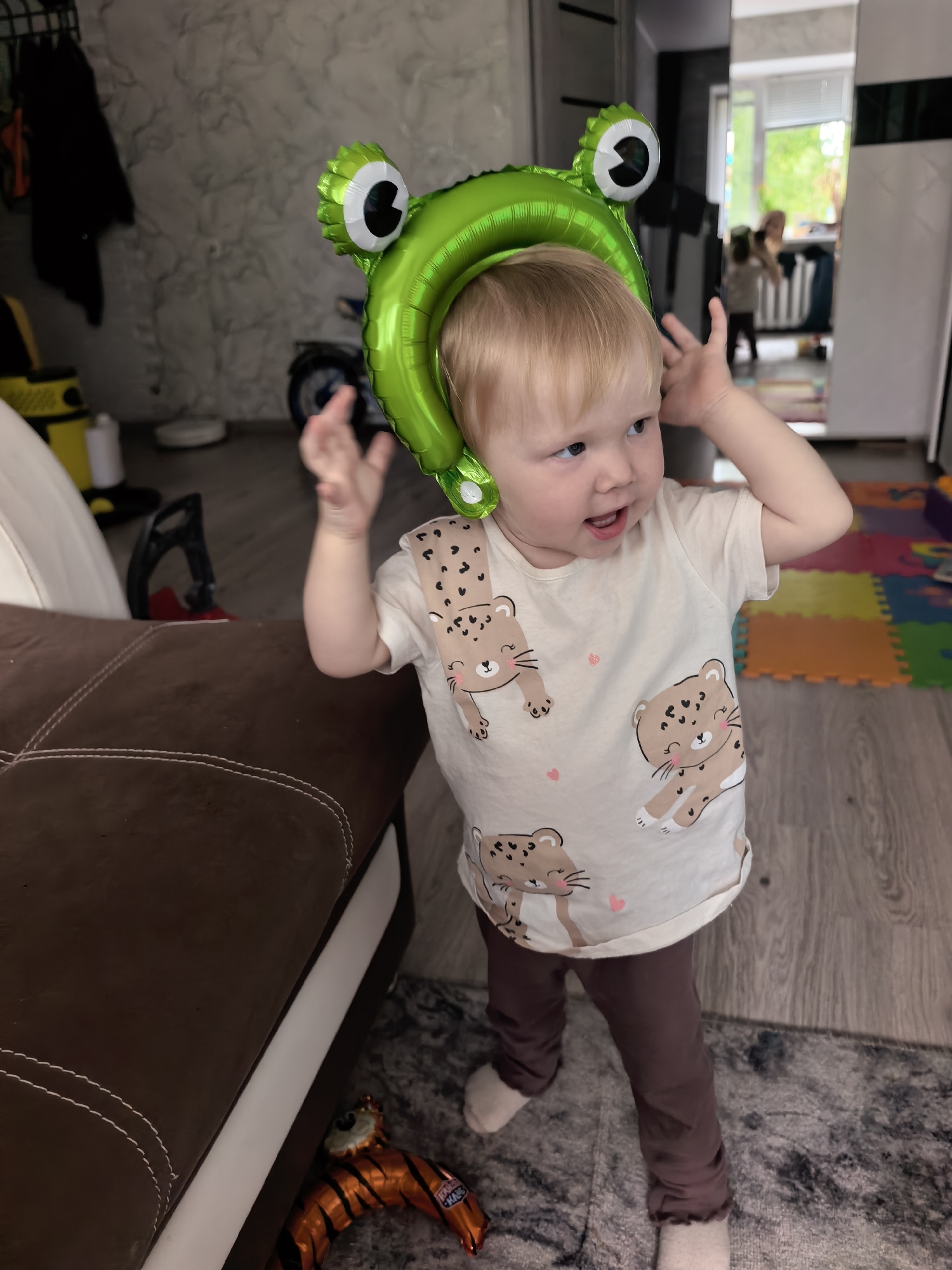 A young child with blonde hair is standing in a room, wearing a light-colored t-shirt with a pattern of cartoon leopards and a frog-shaped balloon on their head. The child is looking to their right with their mouth slightly open and their hands near their head. In the background, there is a brown sofa, a rug, a colorful play mat, and some furniture, including a mirror reflecting a window and other people. A tiger-shaped balloon is on the floor near the sofa. The image is taken indoors. The specific location of Sterlitamak, Russia is not discernible from the visual information present in the image.