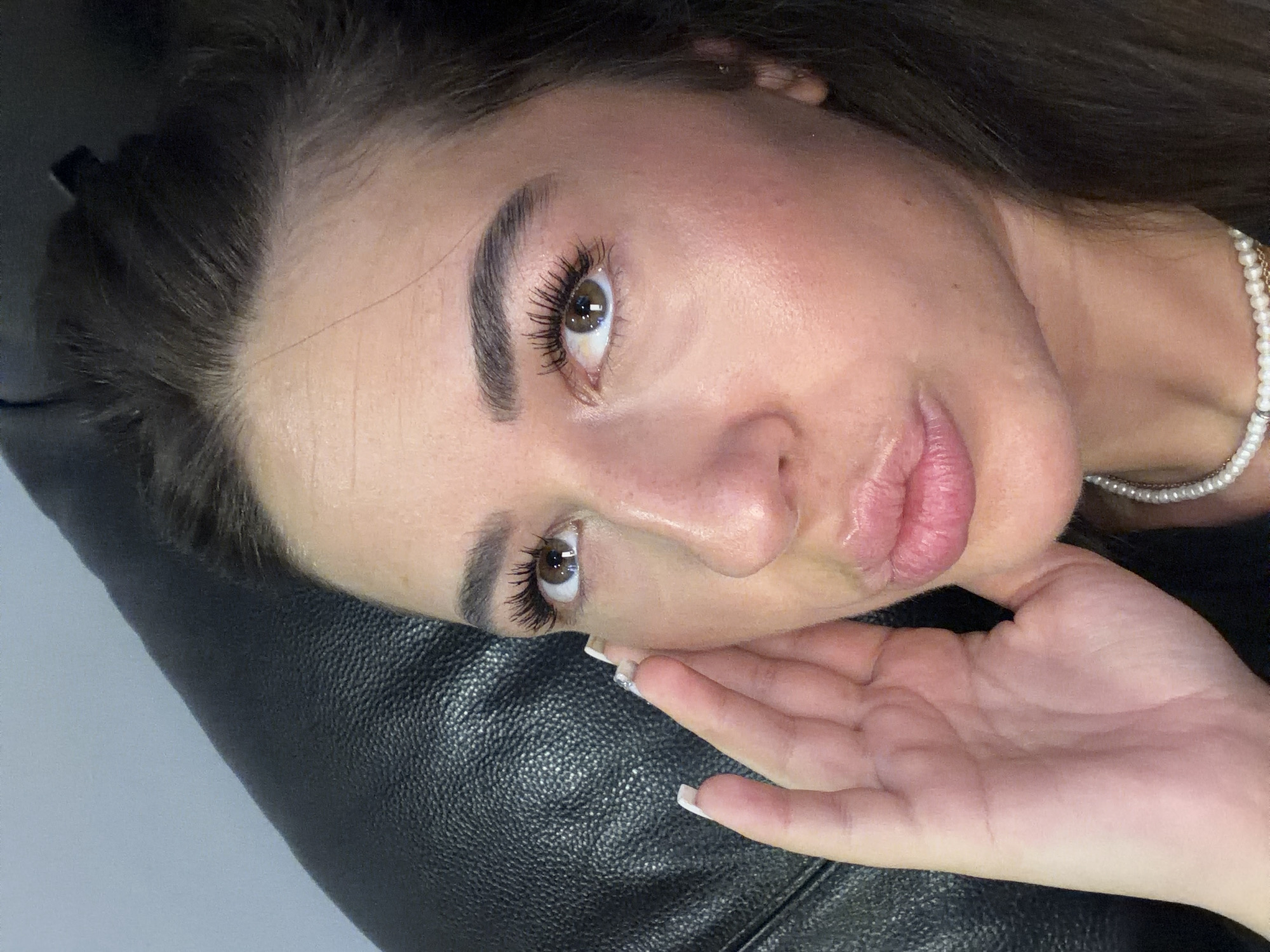 In Novi Iskar, Bulgaria, a close-up portrait captures a young woman against a dark, textured background, likely a leather couch. She is positioned in the upper right quadrant of the frame, her face angled upwards and to the left. Her right hand, with meticulously manicured white nails, is raised to her cheek, fingers gently touching her skin. The woman has dark hair that cascades over her shoulder, and her makeup is evident with pronounced eyelashes and defined eyebrows. Her lips are slightly parted, giving a pouty expression. A strand of pearls is visible around her neck, resting on her black clothing. The lighting highlights the contours of her face, emphasizing her skin's texture and a subtle sheen, suggesting a humid or dewy finish.