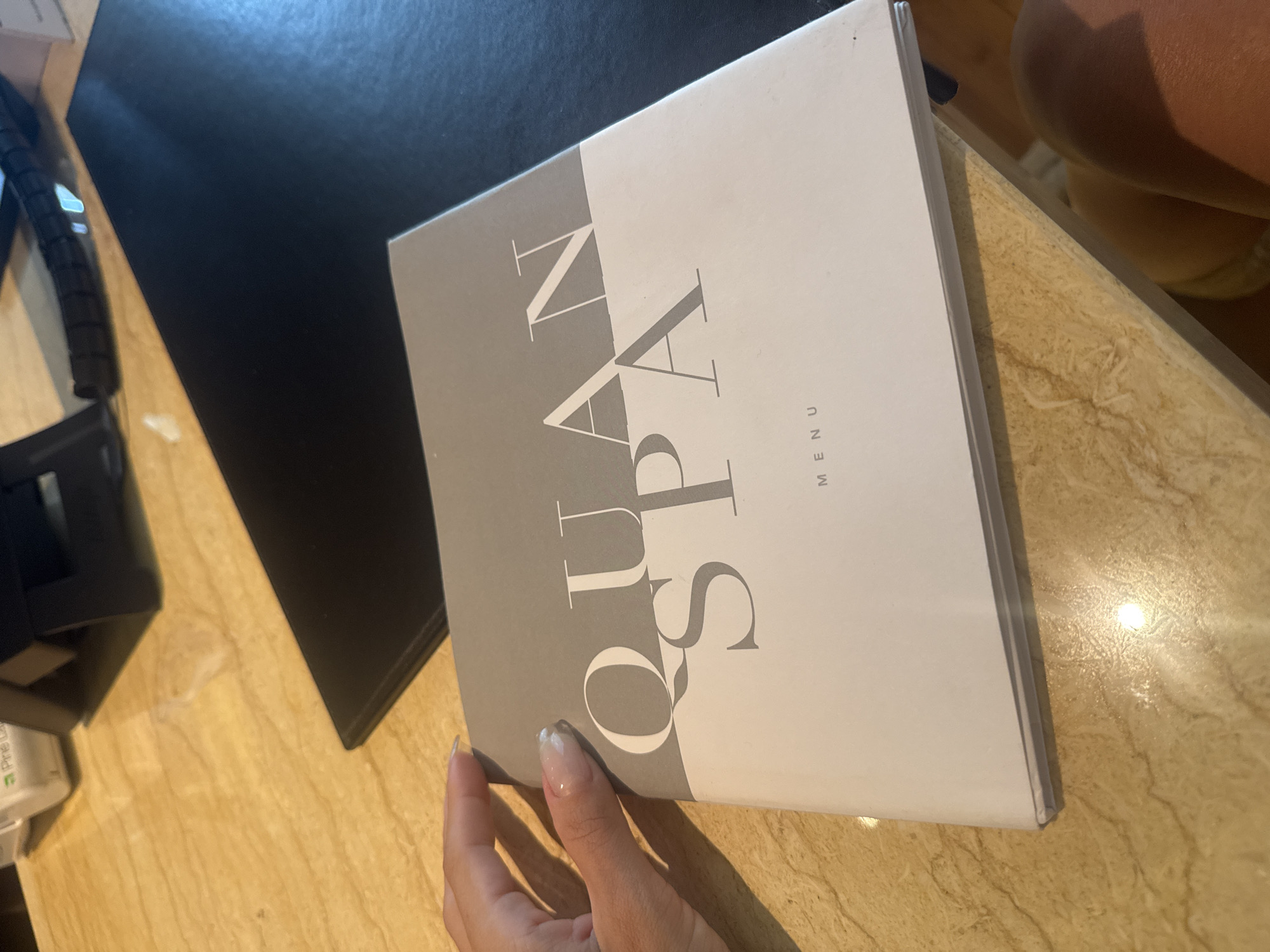 A hand with light-colored nail polish is holding a menu for "QUAN SPA MENU." The menu is a booklet with a grey upper section and a white lower section. The background is a wooden table with a black mat on the right side. The scene is likely indoors in a spa or salon, but there is no specific information to suggest it is in Pune, India.