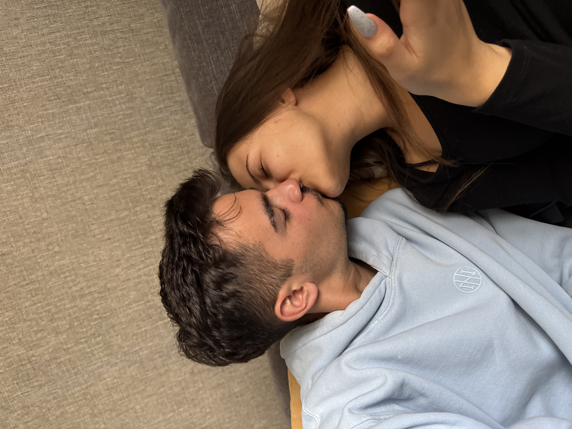 An intimate, diagonally oriented indoor shot captures a man and a woman in a tender kiss. They are lying down, likely on a sofa or bed with a light-colored, textured fabric covering.
The woman, with long, dark brown hair, is positioned above the man, leaning down to kiss him. She is wearing a dark, possibly black, long-sleeved top. Her right hand is visible near her head, featuring long, light gray or light blue painted fingernails. Her eyes are closed, conveying a moment of deep connection.
The man, with dark hair and stubble or a short beard, is lying beneath her, also with his eyes closed. He is wearing a light blue hoodie. On the left chest area of his hoodie, a circular, embroidered logo is visible. It features stylized, intertwined letters, possibly "PNR" or a similar combination, within a circular border.
The lighting appears soft and even, suggesting an indoor setting. The atmosphere is one of affection and closeness. The location context is Sofia, Bulgaria.