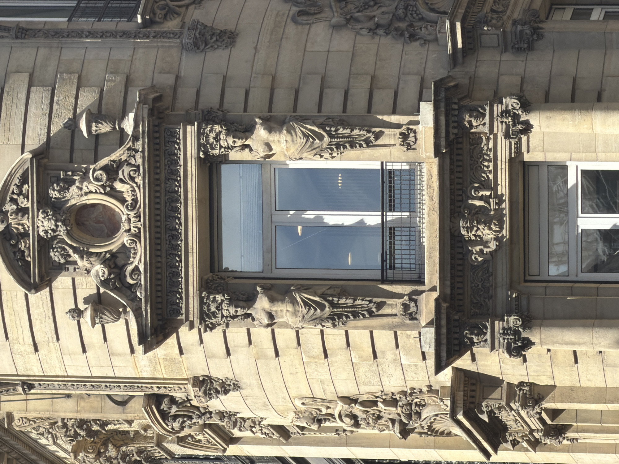 The image shows a building facade in Paris, France. The architecture is ornate and features detailed stone carvings. There are two windows visible, one with a metal grate. Statues of women flank the central window. The facade is light in color.