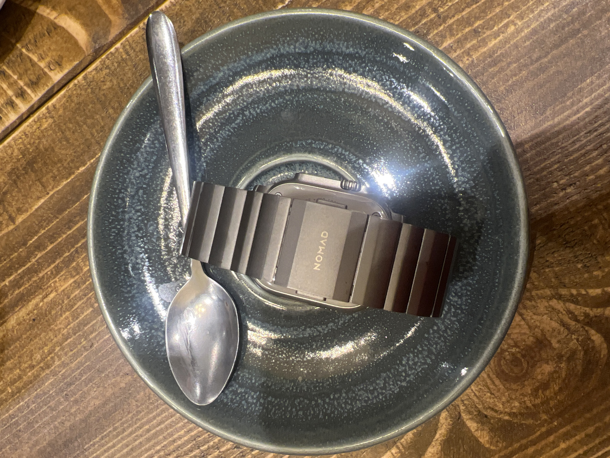 The image shows a grey-blue glazed ceramic bowl resting on a wooden surface. Inside the bowl, there is a silver spoon and a smartwatch with a dark grey metal link band. The band is branded with the word "NOMAD". The spoon lies across the bowl, partially on top of the watch band. The lighting is bright, creating reflections on the spoon and the bowl's surface. The wooden surface appears to be a table with a light brown and dark brown wood grain pattern.
The image does not contain information to determine its location within Mole Valley, United Kingdom.