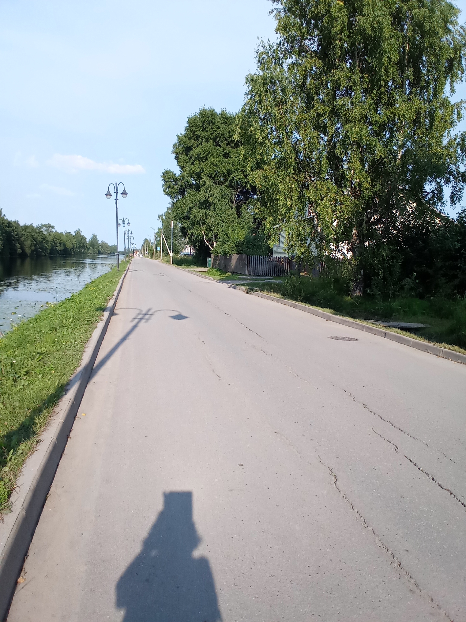 The image depicts a paved road running alongside a calm body of water, likely a canal or river, in the city of Belozersk, Russia. The road is bordered by a grassy embankment on the left, leading down to the water which is partially covered with lily pads. Along the right side of the road, there are several ornate streetlights and lush green trees, with a wooden fence and a building visible in the background. The sky is clear and blue, with a few wispy clouds. The perspective suggests the photo was taken from a standing position, with the shadow of the photographer and their device visible on the road. The road surface shows signs of wear, with cracks and small stones visible. The overall scene conveys a peaceful, somewhat rustic atmosphere characteristic of a summer day in a Russian town.