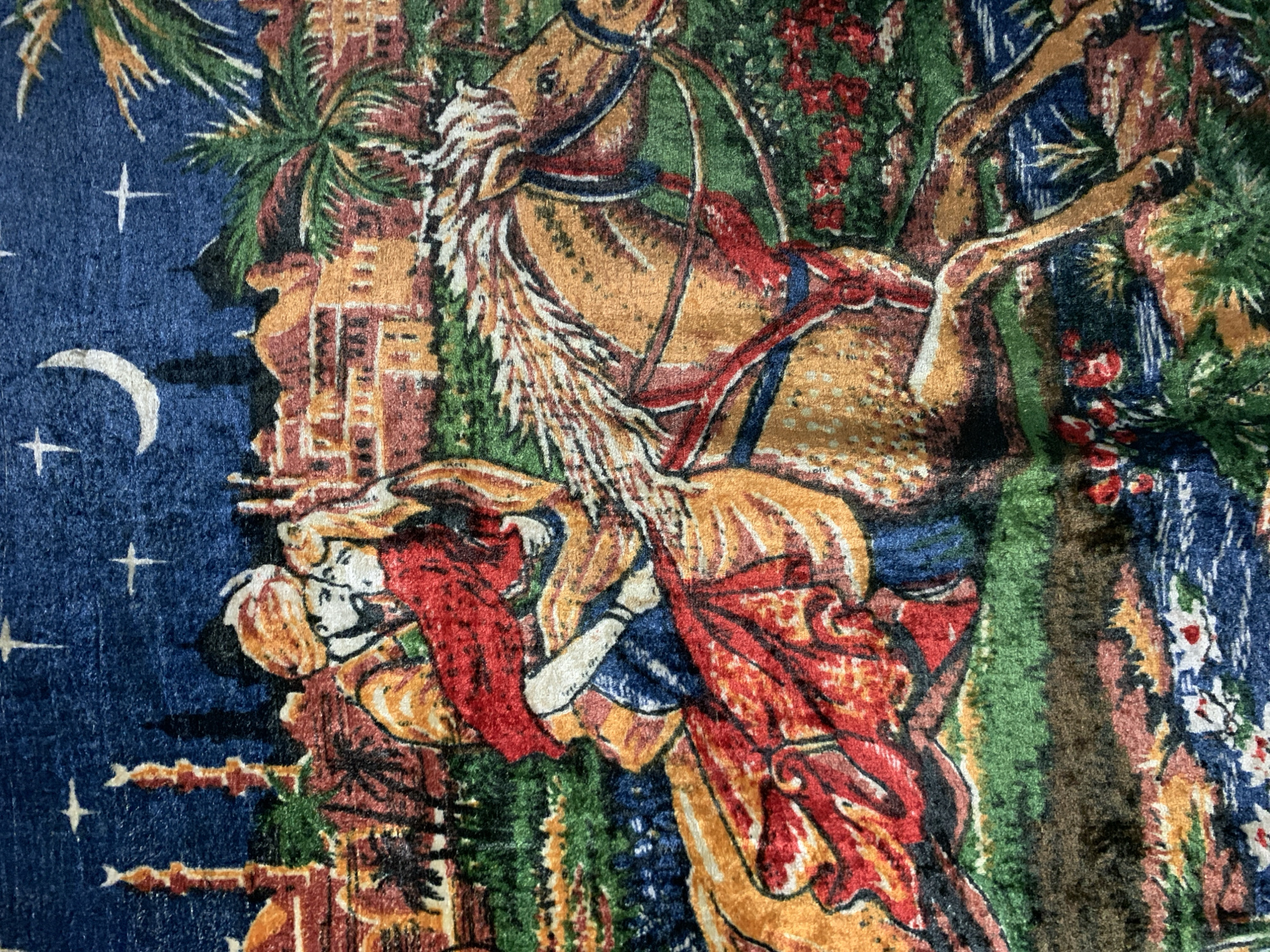 The image depicts a tapestry or fabric with a vibrant, stylized depiction of a romantic scene set against a desert city landscape under a starry night sky with a crescent moon. A man wearing a turban and a woman in a red flowing garment are riding on a horse. The man embraces the woman. The background features architectural elements resembling mosques or minarets, palm trees, and dense foliage in various shades of green and red. The scene is characterized by rich colors and intricate patterns, suggesting an artistic or decorative purpose. The location in Відзаўскі сельскі Савет, Belarus, is not depicted in the image itself; the image is a representation of a scene, not a geographical location.