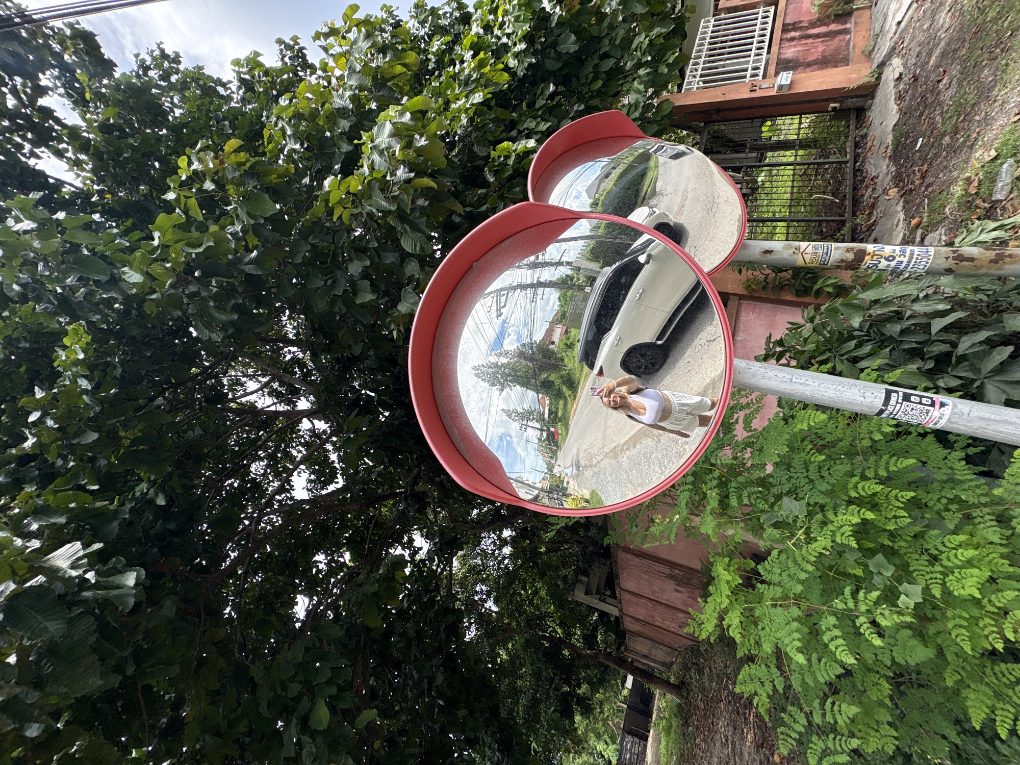 The image shows a convex traffic mirror mounted on a metal pole, reflecting a street scene in Chalong, Thailand. A young woman wearing a white tank top and white shorts is visible in the reflection, holding up her phone to take a selfie. She appears to be smiling.
The mirror reflects a street with a white car, power lines, and buildings in the background. The sky is partly cloudy. Lush green trees and foliage surround the mirror and pole. A pink-colored building is visible to the right of the mirror, with a wooden overhang and a grated fence in front of it.
The time of day appears to be daytime, possibly late morning or early afternoon, given the brightness of the sky and the shadows. The weather seems to be warm and humid, typical of Thailand.
Visible text on the pole includes Thai characters on a sticker, which appears to be a QR code and some sort of advertisement or notice. There is also text on a white sticker below the QR code. The overall impression is a candid snapshot of a moment in a tropical urban environment.