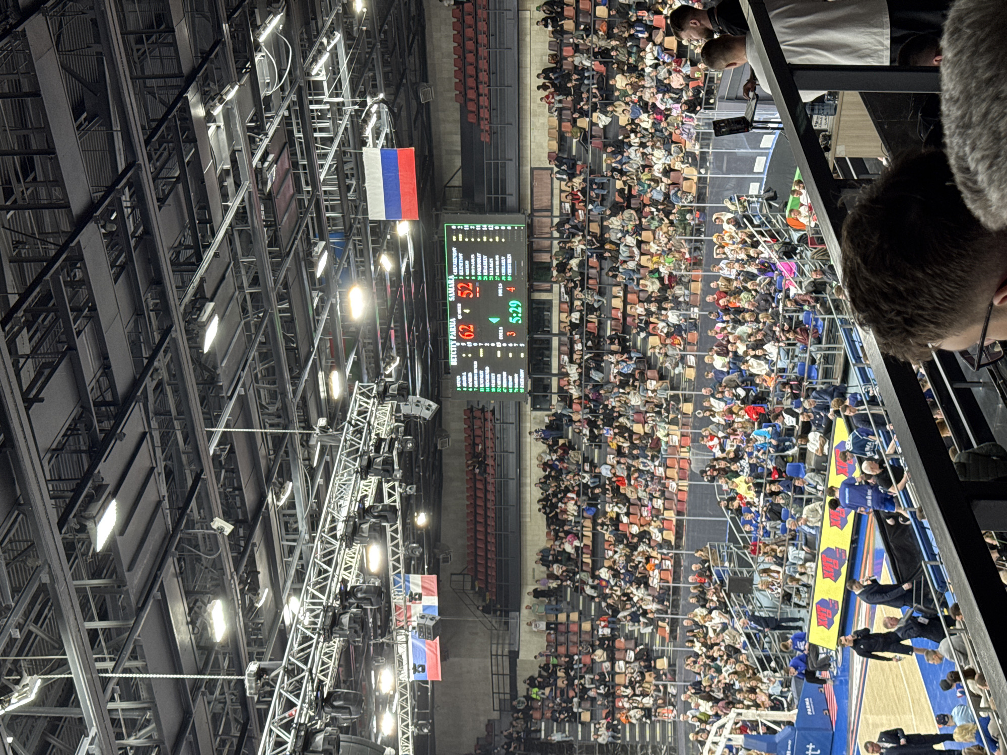 The image captures a basketball game scene within the Molot Sports Hall in Perm, Russia, featuring a large crowd of spectators. The scoreboard shows the game between "Betcity Parma" and "Samara," with the former leading 62 to 52, and the clock displaying "5:29" in the fourth quarter. Russian flags are visible near the scoreboard and lighting rig. People are seated in the stands, and there are individuals on the court sidelines in blue uniforms and suits.