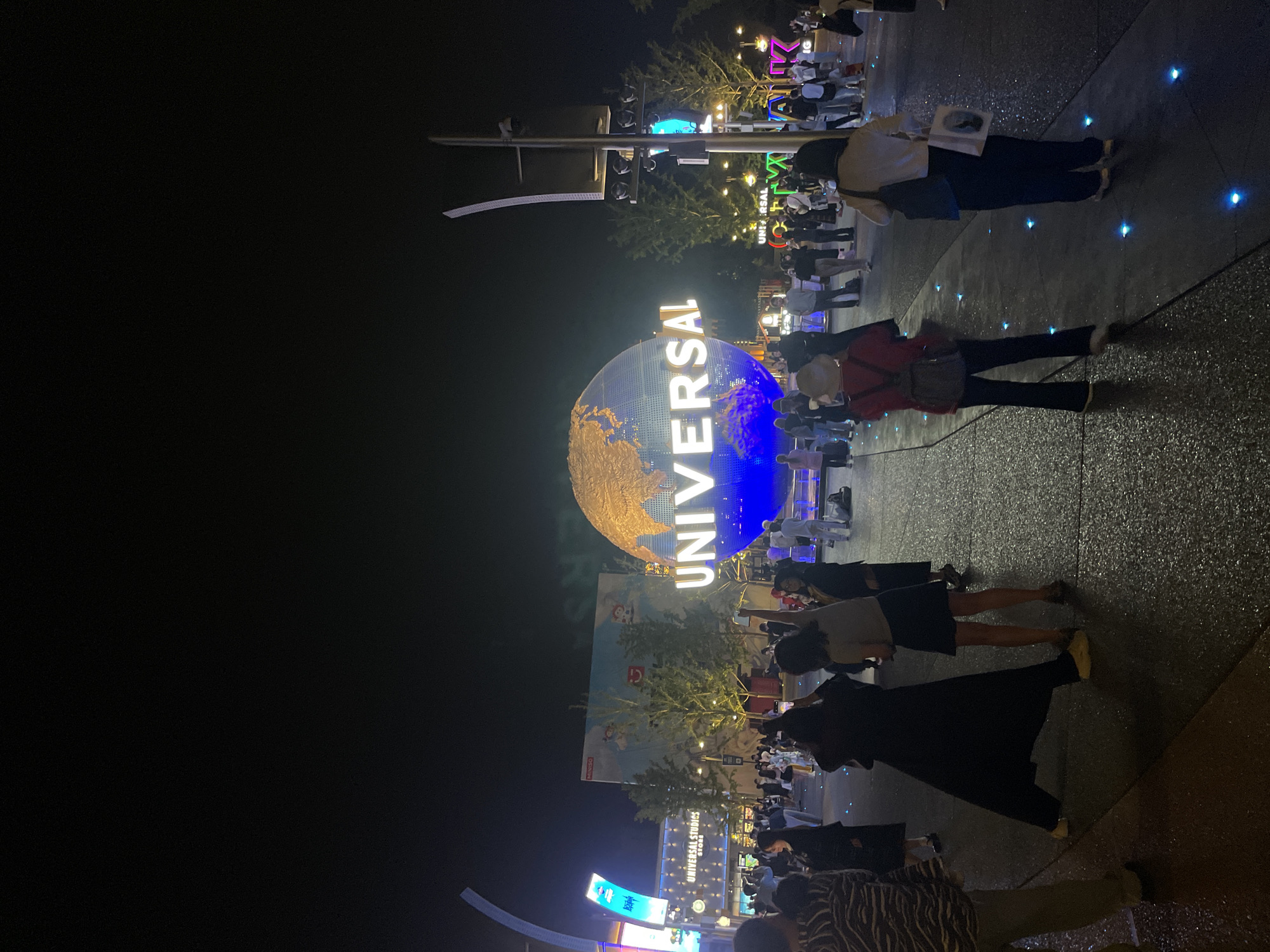 At night in Tongzhou District, China, the Universal Studios globe, emblazoned with the "UNIVERSAL" logo, dominates the scene. The globe is illuminated with gold and blue light. People are walking and standing near the globe. The ground is paved and has embedded blue lights. Lights and signage are visible in the background, along with some trees. The image is shot at an angle.