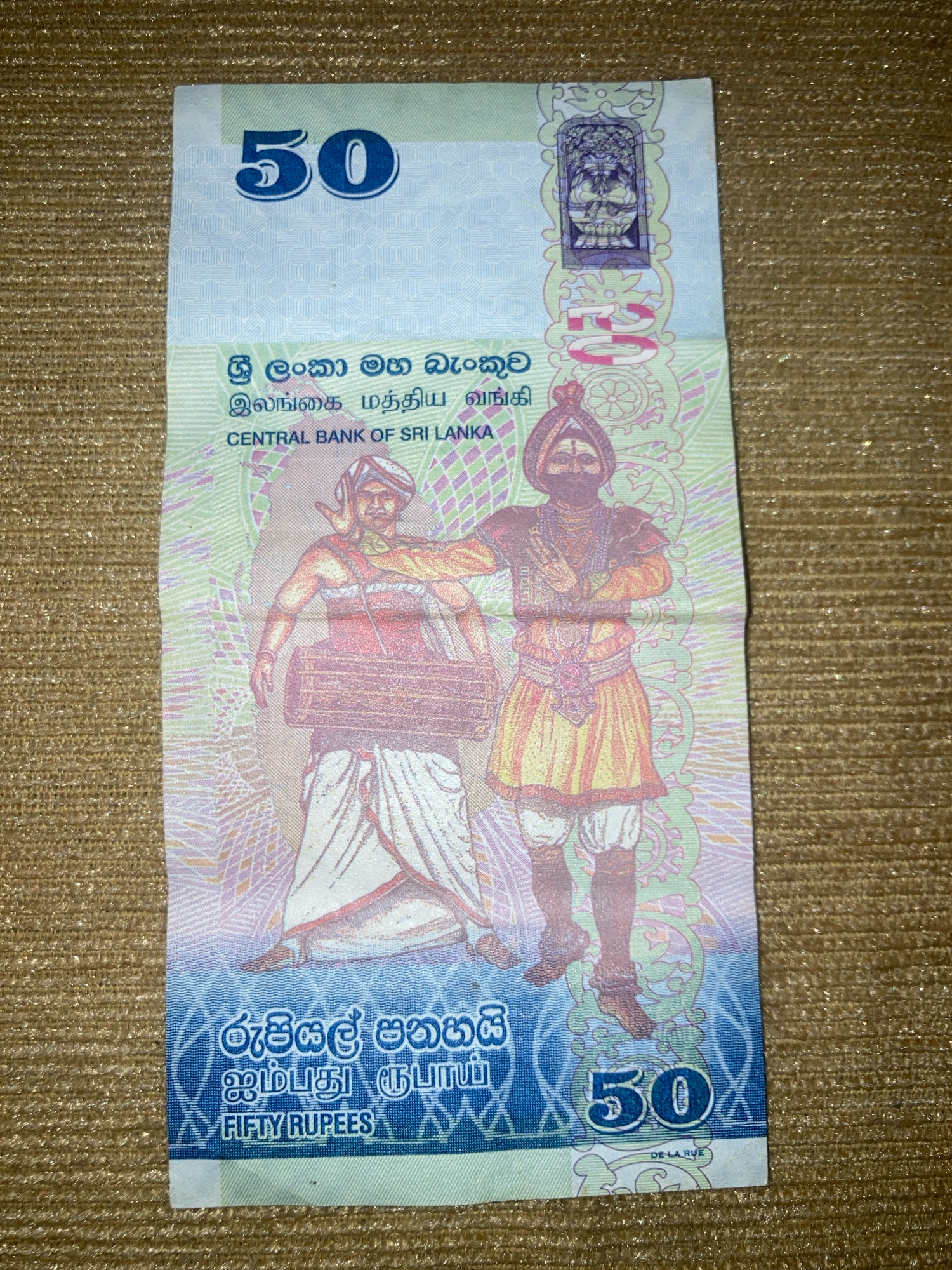 The image is a close-up, top-down shot of a Sri Lankan 50 Rupee banknote. The banknote is predominantly white, blue, and pink, with intricate patterns and illustrations. In the center, two figures in traditional attire are depicted. The figure on the left appears to be playing a drum-like instrument, while the figure on the right stands with arms crossed. Above the figures, the number "50" is prominently displayed in blue. Text in Sinhala, Tamil, and English ("CENTRAL BANK OF SRI LANKA", "FIFTY RUPEES") is also visible. The banknote is laid on a textured, golden surface, possibly a fabric or placemat. The lighting casts a slight shimmer on the surface. The scene is static, with no discernible action taking place.