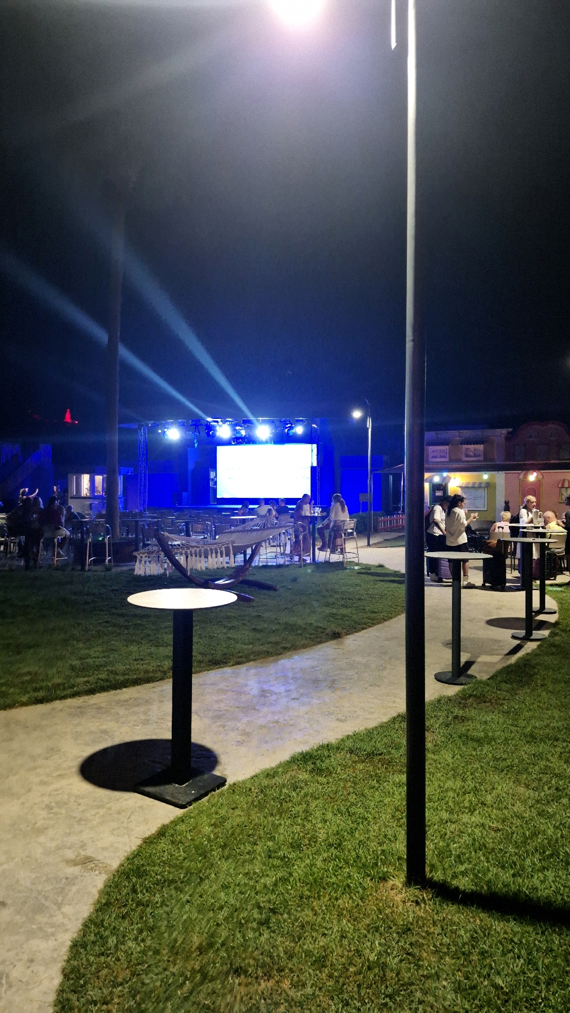 The image depicts an outdoor gathering area at night, possibly a venue or restaurant, with a stage and screen illuminated in the background. The scene is set in the city of Serik, Turkey. In the foreground, a paved pathway curves through a grassy area where several small, round tables with dark bases and white tops are placed. A prominent black lamppost stands to the right, casting a shadow on the path. Further back, near the stage, there are more tables and chairs, some occupied by people. A hammock is suspended between two poles. The stage is bathed in blue light, with bright white spotlights directed upwards, creating beams that cut through the night sky. Small structures resembling shops or booths are visible behind the stage area. The overall atmosphere suggests a relaxed evening event.