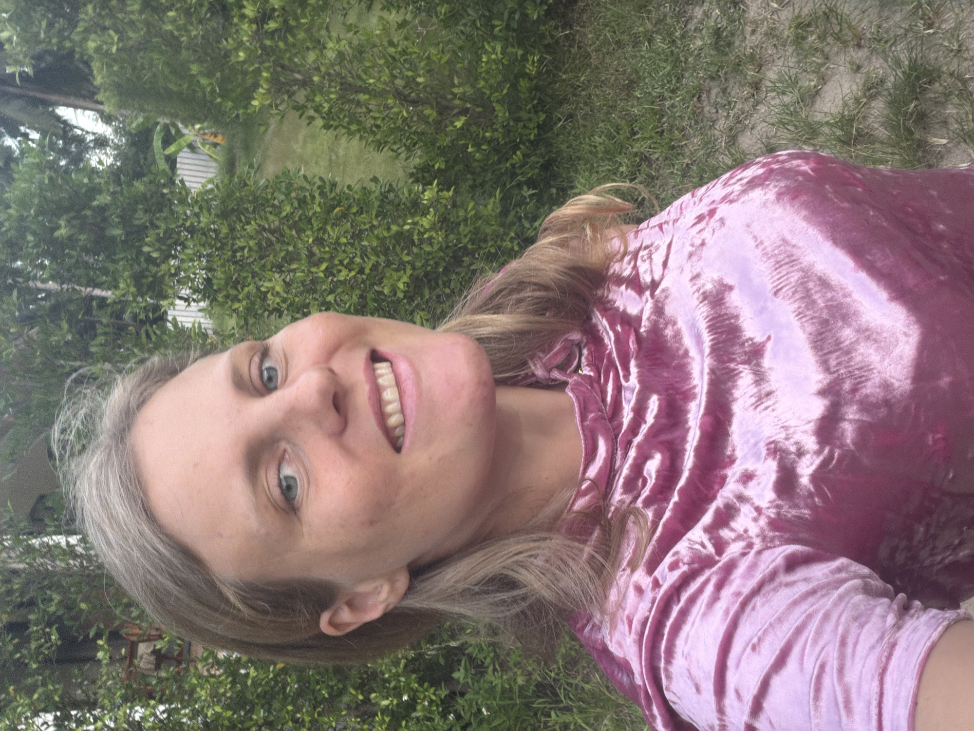 A blonde woman with light blue eyes is pictured in an outdoor setting, smiling. She has shoulder-length, wavy blonde hair and is wearing a shiny pink, long-sleeved top. The background consists of lush green foliage, including trees and grass, suggesting a tropical environment. The location is identified as Koh Phangan Sub-district, Thailand.