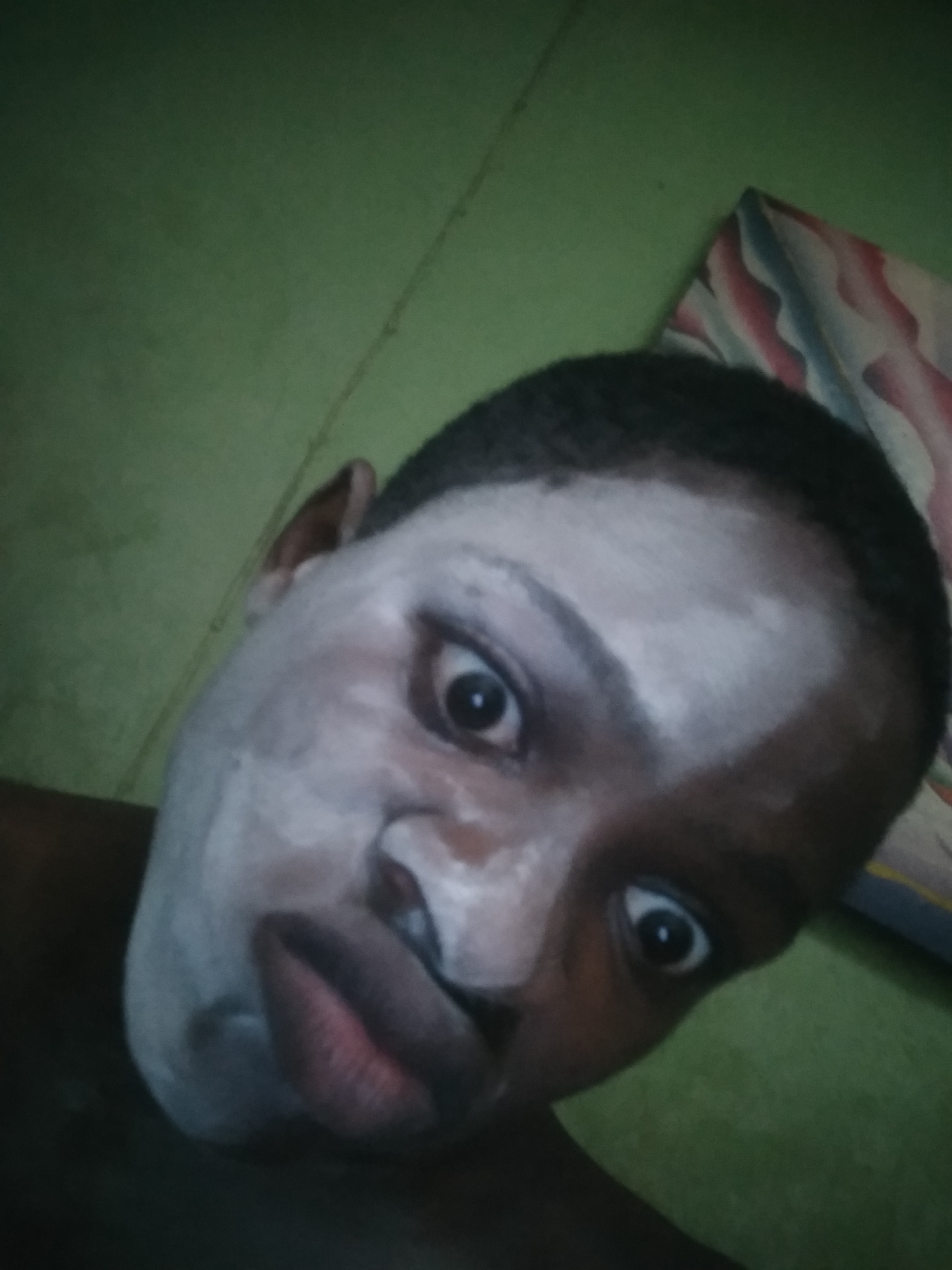 A close-up, selfie-style photograph shows a person with dark skin, a shaved head, and a light-colored substance applied to their face. The person's eyes are wide open and staring directly at the camera. The background includes a light green wall and a patterned object, possibly a pillow. The location, Shangisha, Nigeria, is not evident in the image and cannot be determined from the visual content alone.