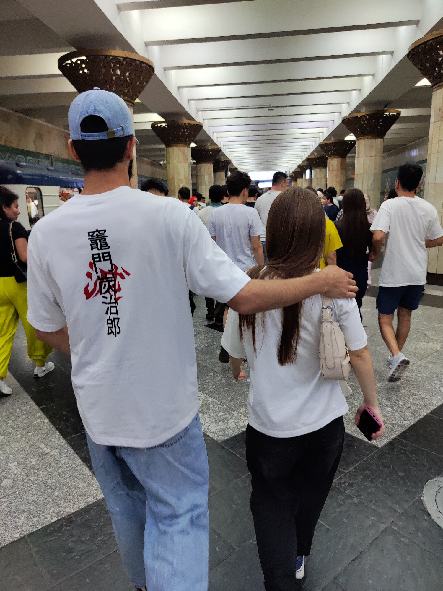 In Tashkent, Uzbekistan, a man wearing a light blue baseball cap and a white t-shirt with Japanese calligraphy on the back has his arm around a woman's shoulders. The woman is wearing a white t-shirt and black pants, and carrying a light beige purse. They are walking away from the camera through a well-lit subway station, characterized by ornate columns and a tiled floor. Other pedestrians are visible in the background, moving in the same direction.