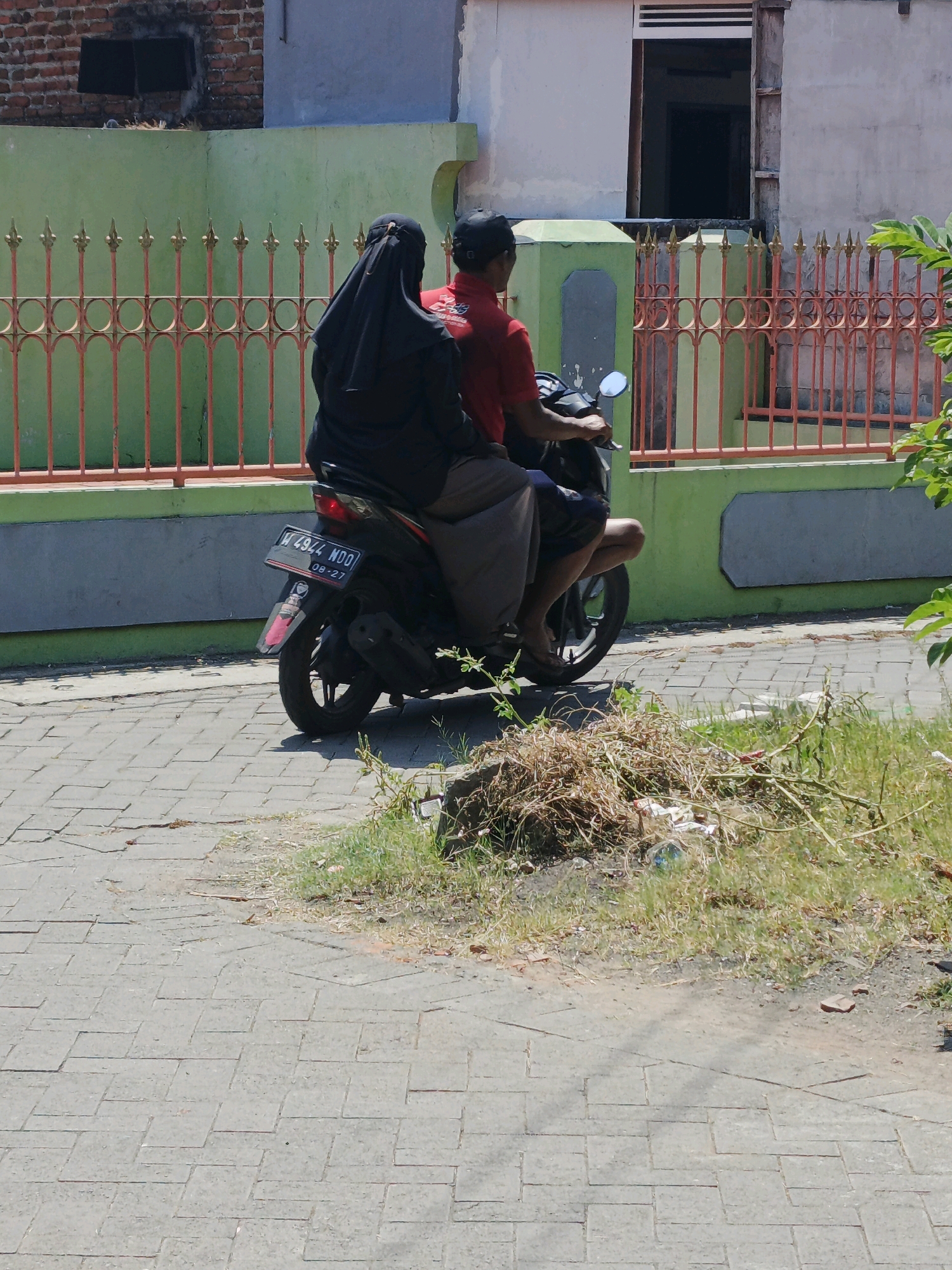In Ketegan, Indonesia, two individuals are riding on a scooter on a paved road. The person in the back, wearing a black hijab and dark clothing, is seated behind the driver. The driver, wearing a red shirt and a dark cap, is operating the scooter. A motorcycle license plate visible on the back of the scooter reads "W 4944 WDO" with an expiration date of "08-27". The background features a green wall and a red-barred fence, with a building structure behind it. The environment appears to be outdoors, with dry grass and debris along the side of the road in the foreground.