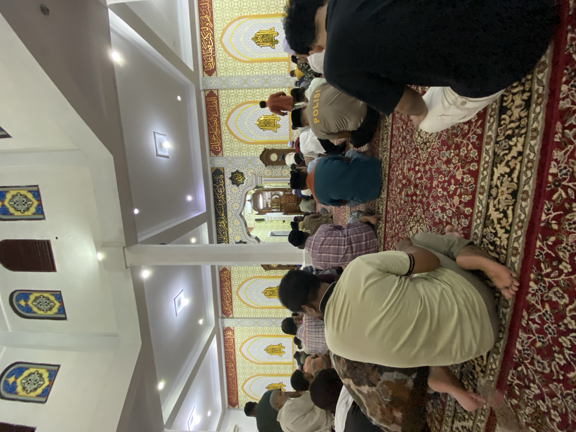 In a mosque in Lhokseumawe, Indonesia, a group of men are gathered for prayer, seated on a red carpet adorned with an intricate floral pattern. The walls of the mosque are decorated with yellow and white Islamic calligraphy and geometric designs, interspersed with arched motifs. Above, the ceiling is a light color with recessed lighting fixtures. The men are dressed in various casual clothing, including shirts, t-shirts, and some wearing caps. One individual in the foreground wears a beige t-shirt with the word "POLISI" (Police) printed on the back. Some individuals are partially obscured by others or by their seating position. The atmosphere appears to be one of communal worship and quiet reverence.