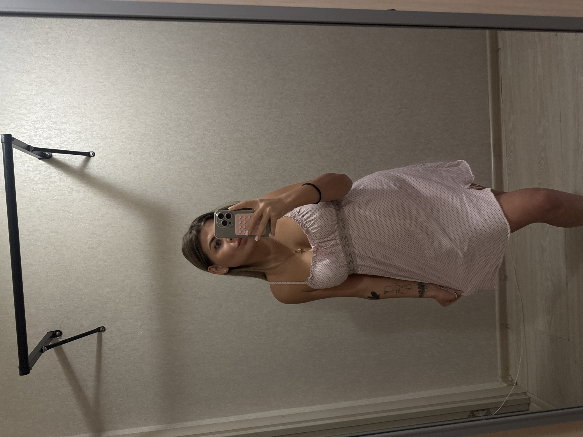 A fair-skinned woman is taking a selfie in a mirror. She is wearing a light pink, polka-dotted dress with lace trim. She is holding a smartphone with her left hand. A black bracelet is on her left wrist, and there is a tattoo on her left forearm. A gold necklace is visible on her chest.
The mirror is framed in grey and reflects a wall and a clothes-drying rack. The wall is a light grey color. The background also reveals a wall made of wood-like panels. The reflection suggests the location is indoors, possibly a closet. There is no explicit information to confirm the location as Chaikovsky, Russia.