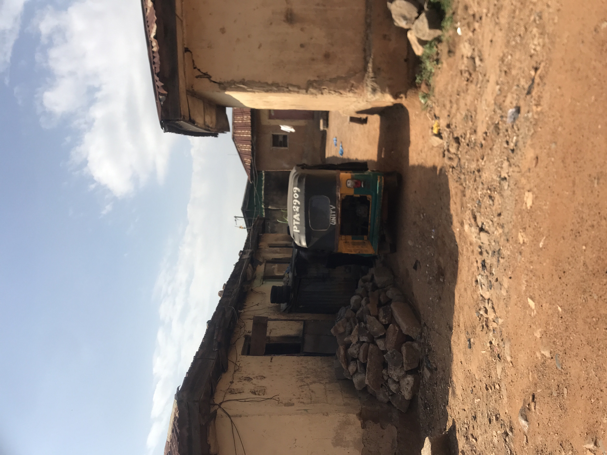 The image depicts a scene in a dusty, urban environment, likely within the city of Rafin Par, Nigeria. A colorful auto-rickshaw, identified by the license plate "PTA 2909" and the word "UNITY" on its rear, is parked on a dirt road. The auto-rickshaw is positioned between two weathered, single-story buildings. The building on the left has a corrugated metal roof and a rough, cream-colored exterior wall with visible cracks and some discoloration. A pile of large, rough stones is stacked against this building. The building on the right, also with a similar roof, has a light brown, textured facade, showing cracks and signs of age. In the background, more structures with corrugated metal roofs are visible under a bright, partly cloudy sky. The ground is composed of reddish-brown dirt and scattered debris. There are no people visible in the scene.