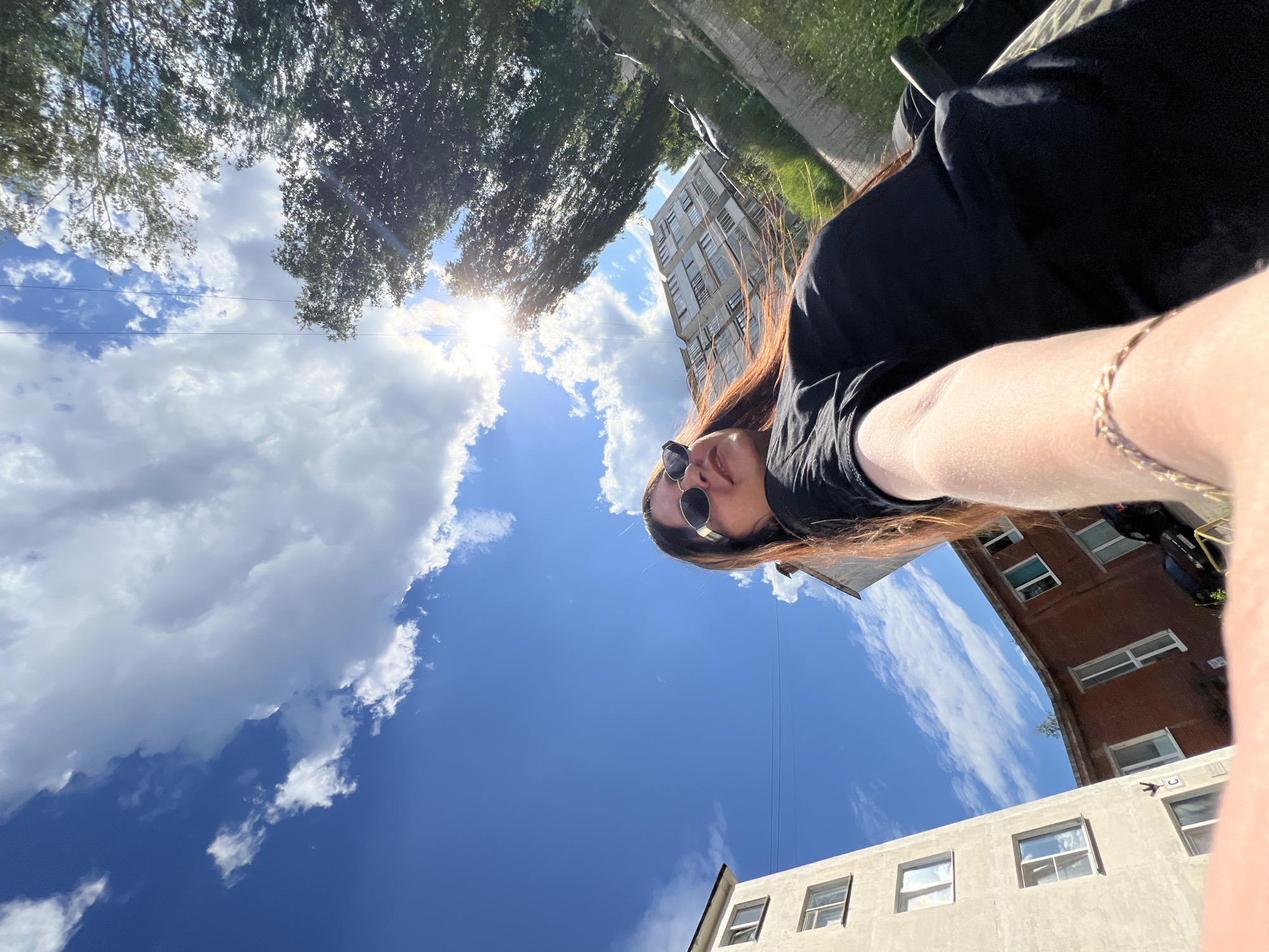 A low-angle, wide shot captures a young woman from the chest up, taking a selfie outdoors on a sunny day in Noviy, Russia. She is wearing sunglasses and a black t-shirt, with her auburn hair falling over her shoulders. The background features a bright blue sky with fluffy white clouds, a tall apartment building, and a smaller brick building. Trees line the right side of the frame, with sunlight filtering through their leaves. The scene appears to be set in a residential area, possibly a courtyard, given the presence of buildings and greenery.