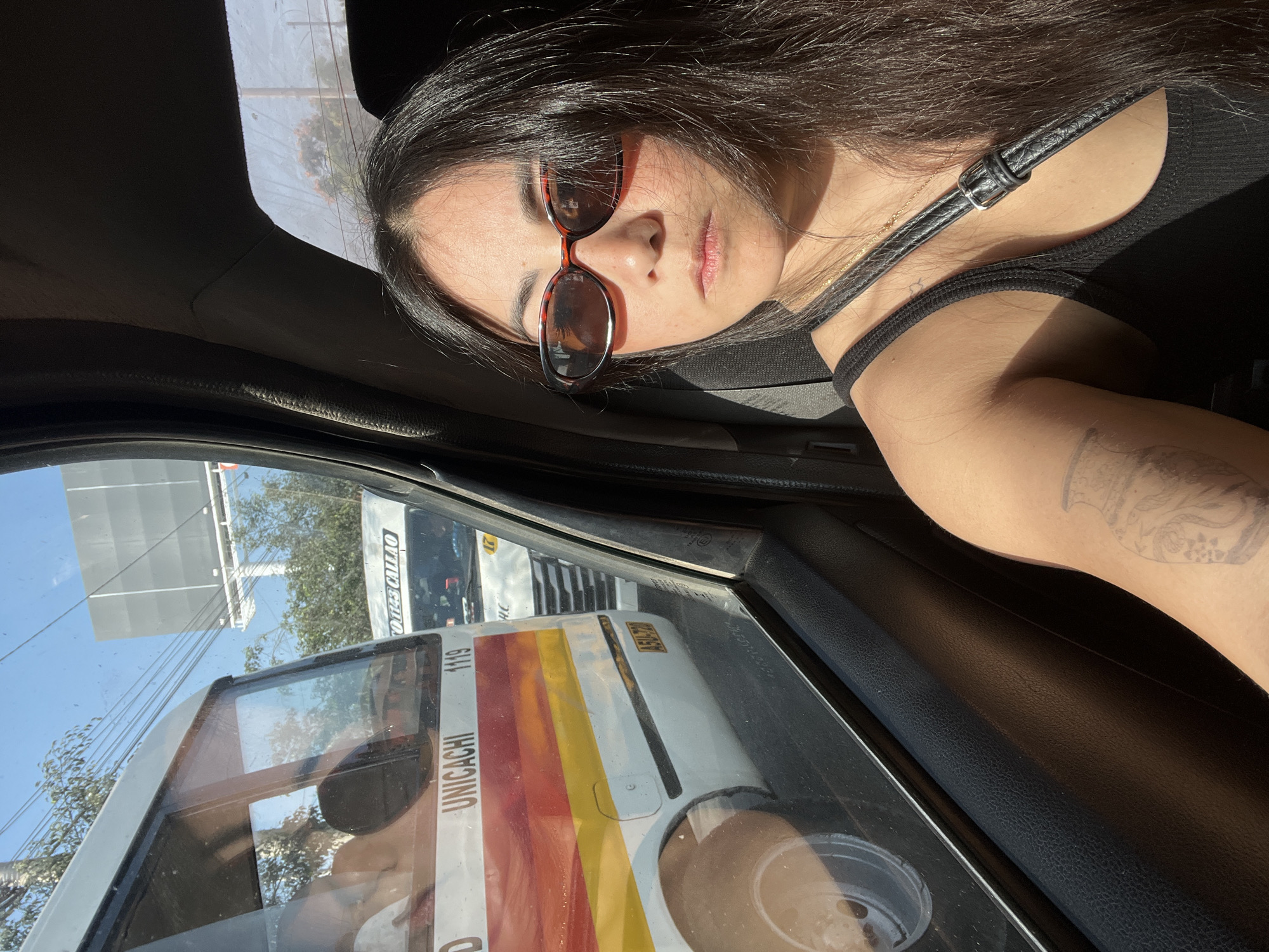 A woman wearing a black tank top and sunglasses is in the passenger seat of a car. She is looking to her right, out the window. Outside the car, a white bus with red and yellow stripes is visible. The bus has the word "UNICACHI" and the number "1119" on its side. Another white vehicle is behind the bus. The scene appears to be in San Borja, Peru, based on the context of the provided information. The sun is shining, casting a shadow on the woman's face. There is a tattoo on her left arm.