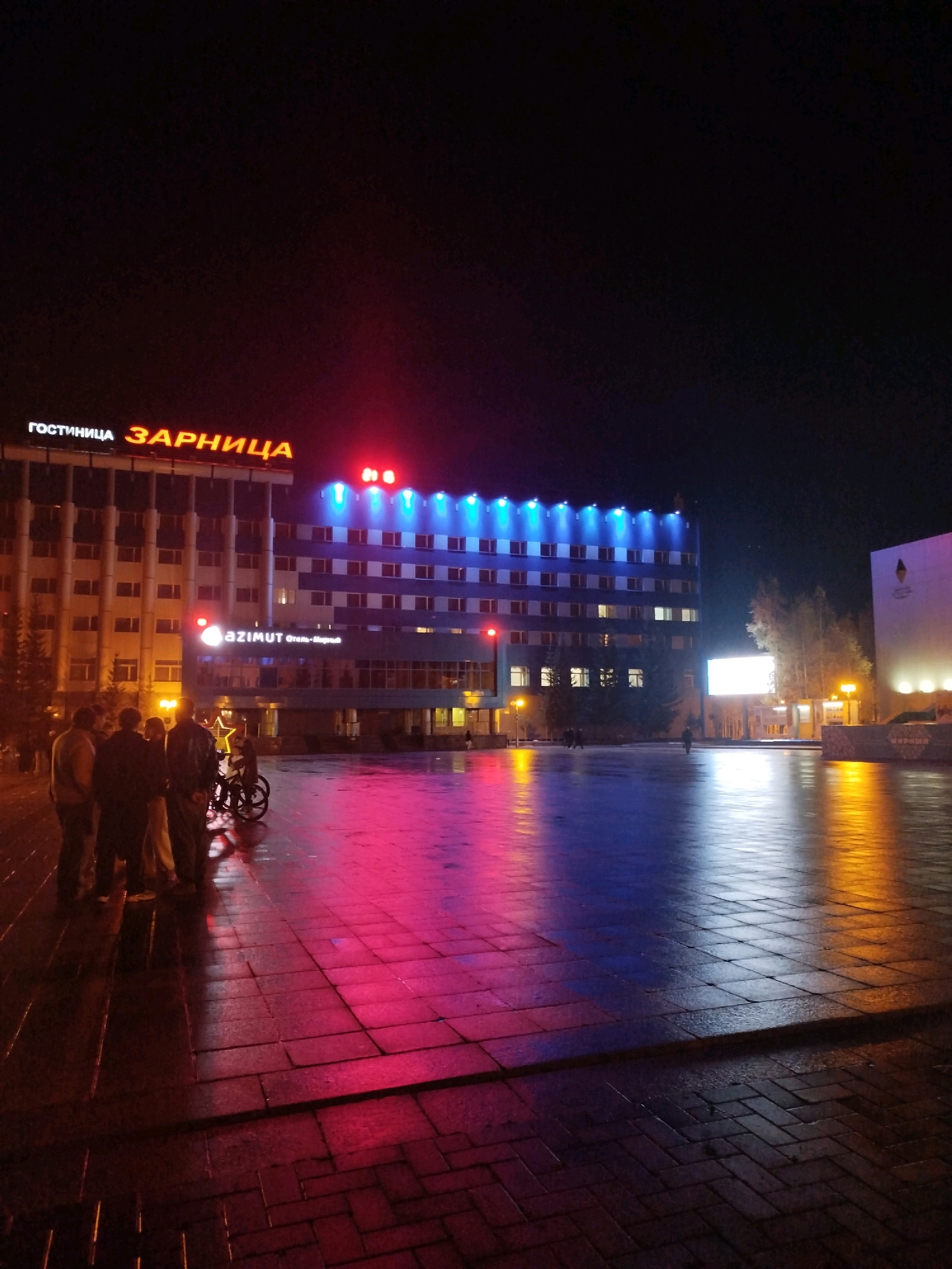A nighttime, eye-level shot captures a wet, paved plaza in Mirny, Russia, reflecting vibrant neon lights from surrounding buildings. The central focus is a modern, multi-story hotel with a sign reading "ГОСТИНИЦА ЗАРНИЦА" (Hotel Zarnitsa) illuminated in orange neon. Below this, a circular "AZIMUT" logo with Russian text is visible. The hotel's facade is lit by rows of blue lights, and its windows glow with a warm yellow light. To the right, another building with a large, illuminated screen and a geometric logo is present. In the foreground, a group of five individuals stands clustered together on the left side of the plaza, facing the hotel. One person is near a bicycle. The wet pavement creates colorful reflections of the building lights, casting pink, blue, and yellow hues across the ground. The atmosphere is urban and illuminated by artificial light.