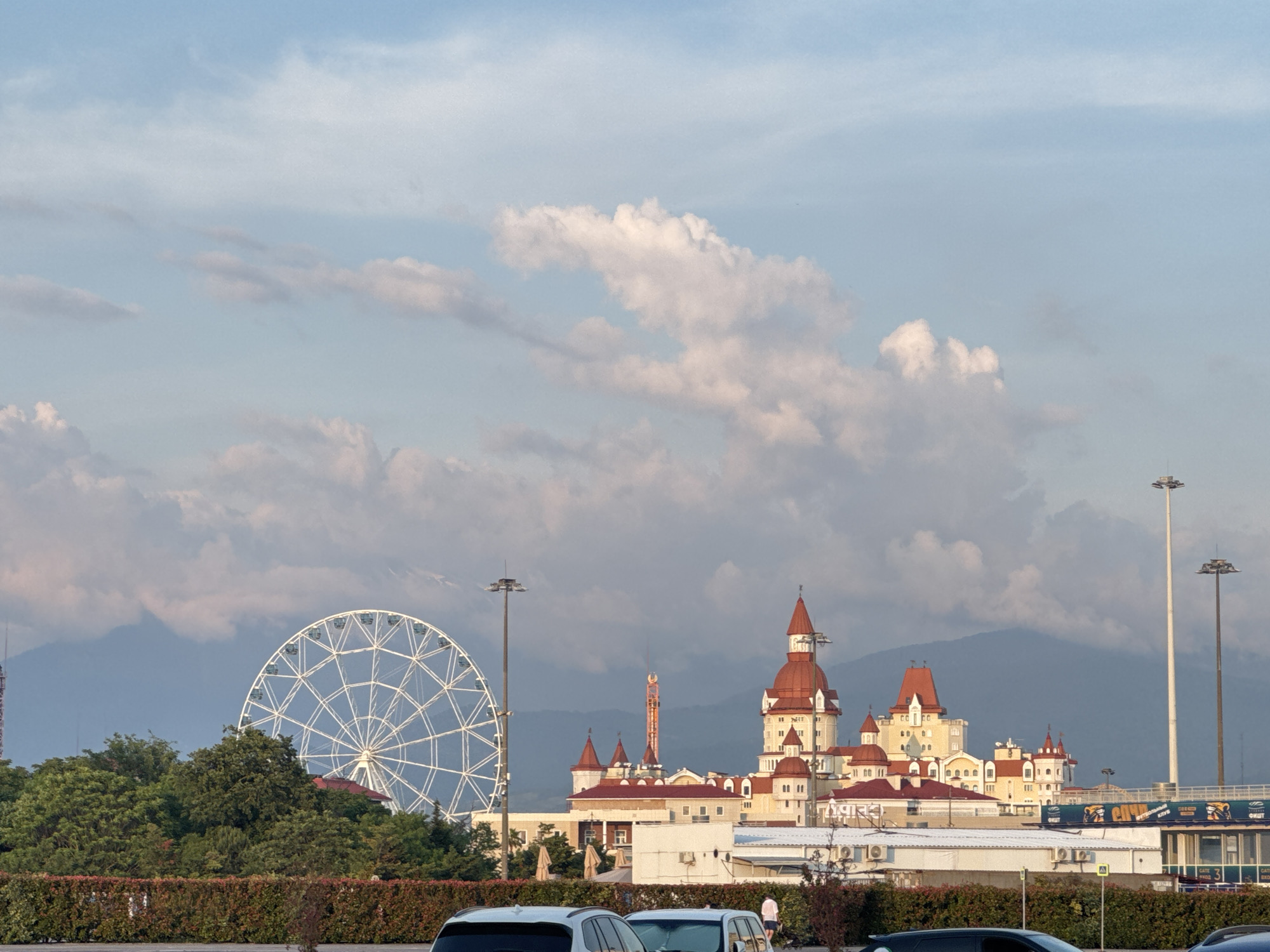 The image depicts a daytime scene in Sirius, Russia, showcasing a complex of buildings with a distinctive fairytale-like architecture, characterized by red roofs and turrets, set against a backdrop of mountains. To the left, a large white Ferris wheel stands prominently, surrounded by lush green trees. In the foreground, a row of bushes and hedges separates the area from a parking lot where several cars are visible. The sky is a pale blue, adorned with scattered white clouds. The overall impression is one of a tourist or entertainment complex.