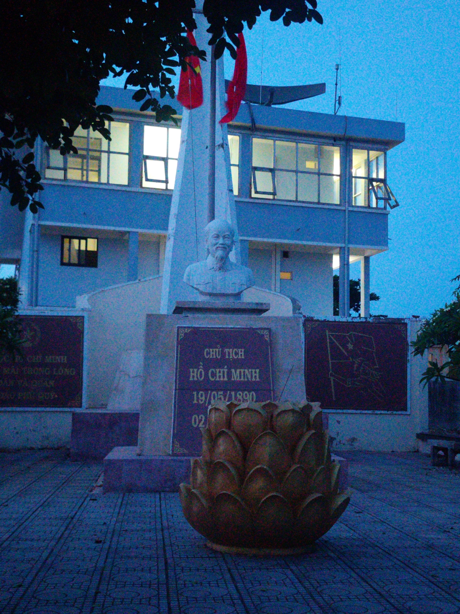 The image shows a statue of Ho Chi Minh, a Vietnamese revolutionary leader, located in the city of Phú Quý Special Administrative Region, Vietnam. The statue is a bust mounted on a pedestal with an inscription that reads "CHỦ TỊCH HỒ CHÍ MINH 19/05/1890." Two red Vietnamese flags are positioned behind the monument. In front of the monument is a golden lotus-shaped sculpture. The monument is located in front of a two-story building.