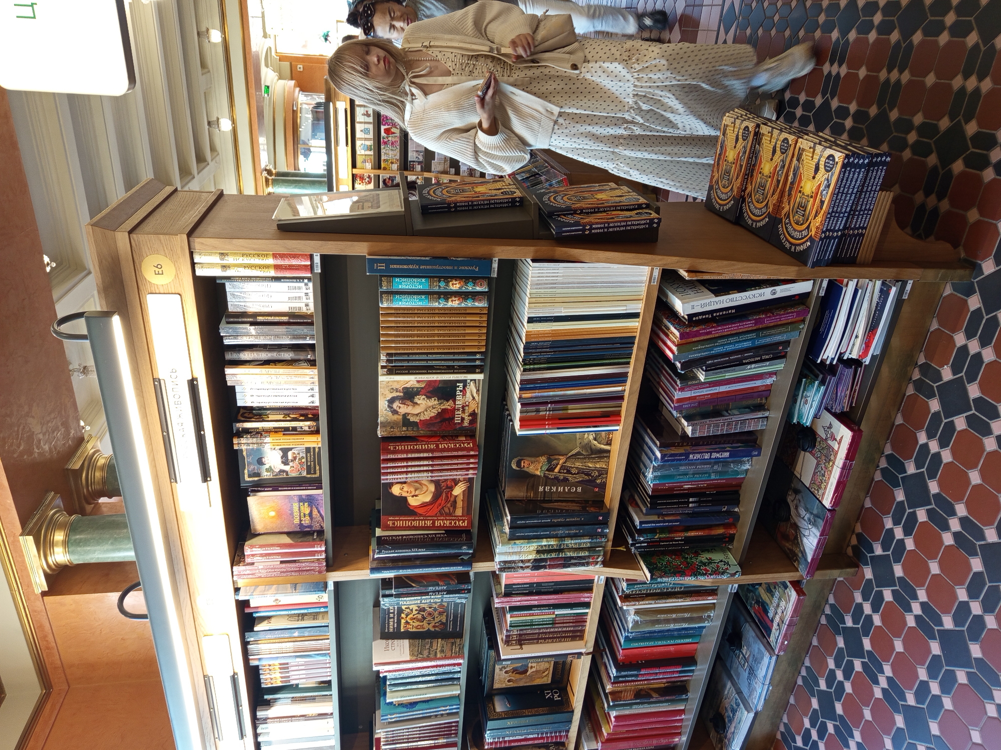 This media file shows an interior view of a bookstore in Saint Petersburg, Russia. The scene is filled with bookshelves packed with books. In the foreground, a wooden bookshelf displays various titles, including what appear to be art and history books. Some books are arranged in stacks, while others are neatly lined up on the shelves.
In the background, two women are partially visible, appearing to browse the books. One woman in the foreground has blonde hair and is wearing a cream-colored cardigan and a polka-dot dress. She is holding what looks like a mobile phone or a small notebook. The other woman, behind her, is wearing glasses and a lighter-colored top. They seem to be casually shopping in the bookstore.
The setting includes architectural details like columns and ornate flooring with a distinctive hexagonal tile pattern in shades of brown, grey, and black. The lighting suggests it could be daytime, possibly late afternoon given the warm tones of the light. The overall atmosphere appears calm and focused on the books and the shopping experience.
Visible text includes Russian titles on the book spines. Some titles that can be partially read include "Русское искусство," "Русская живопись," and potentially titles related to Saint Petersburg, indicated by "Петербурга" on some book covers in the foreground. The label "E6" is also visible on the bookshelf.