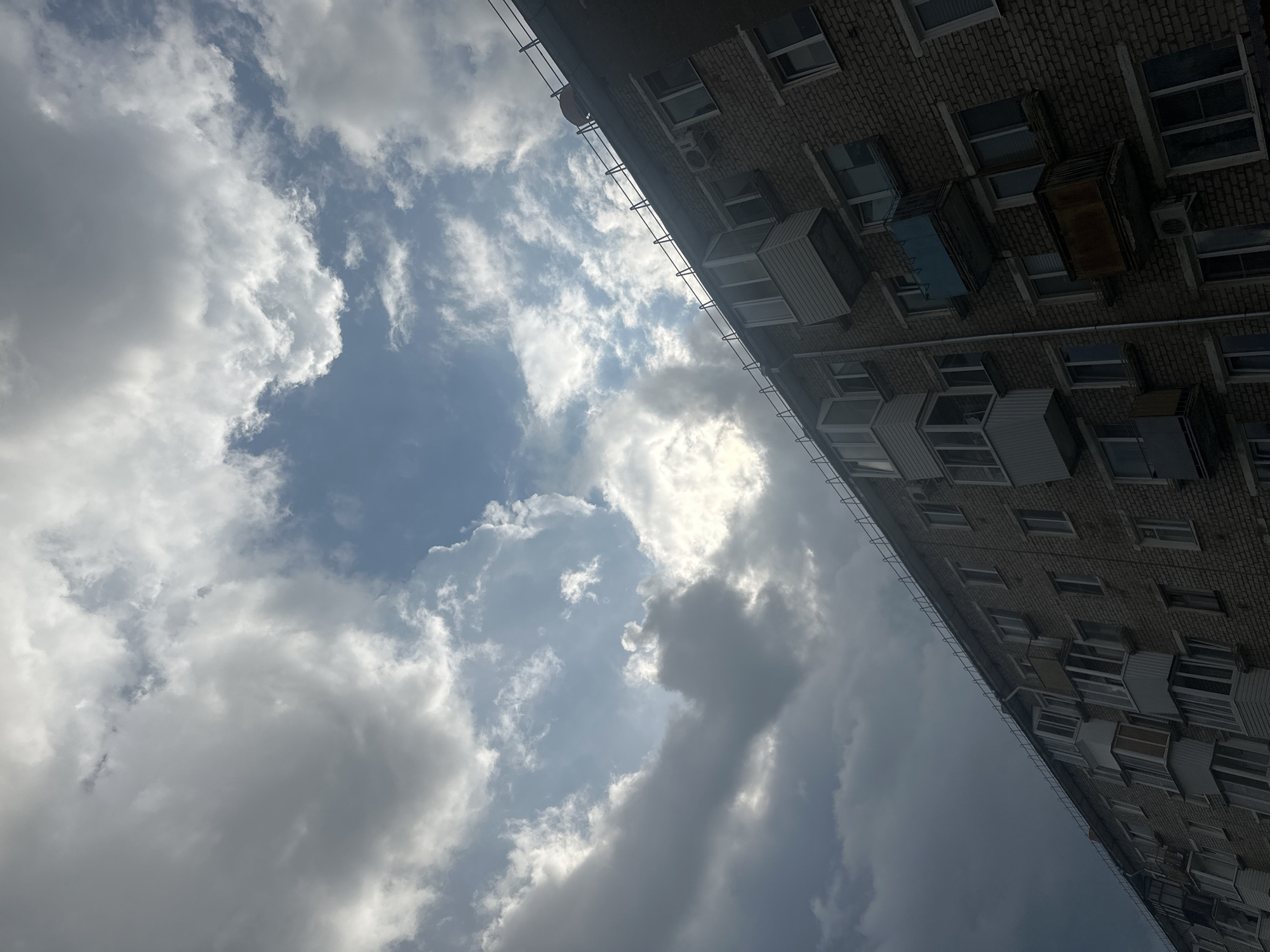 The image captures an upward-angled view of a brick apartment building against a sky filled with dramatic, partly cloudy formations. The building, which appears to be located in Chaikovsky, Russia, shows multiple stories with numerous windows and balconies. Some balconies are enclosed, while others are open. The brickwork is a common reddish-brown. The sky displays a mix of dark, heavy clouds and patches of bright blue, with sunlight breaking through in some areas, creating a contrast between light and shadow. No people are visible in the frame. The scene suggests an overcast or changing weather condition.