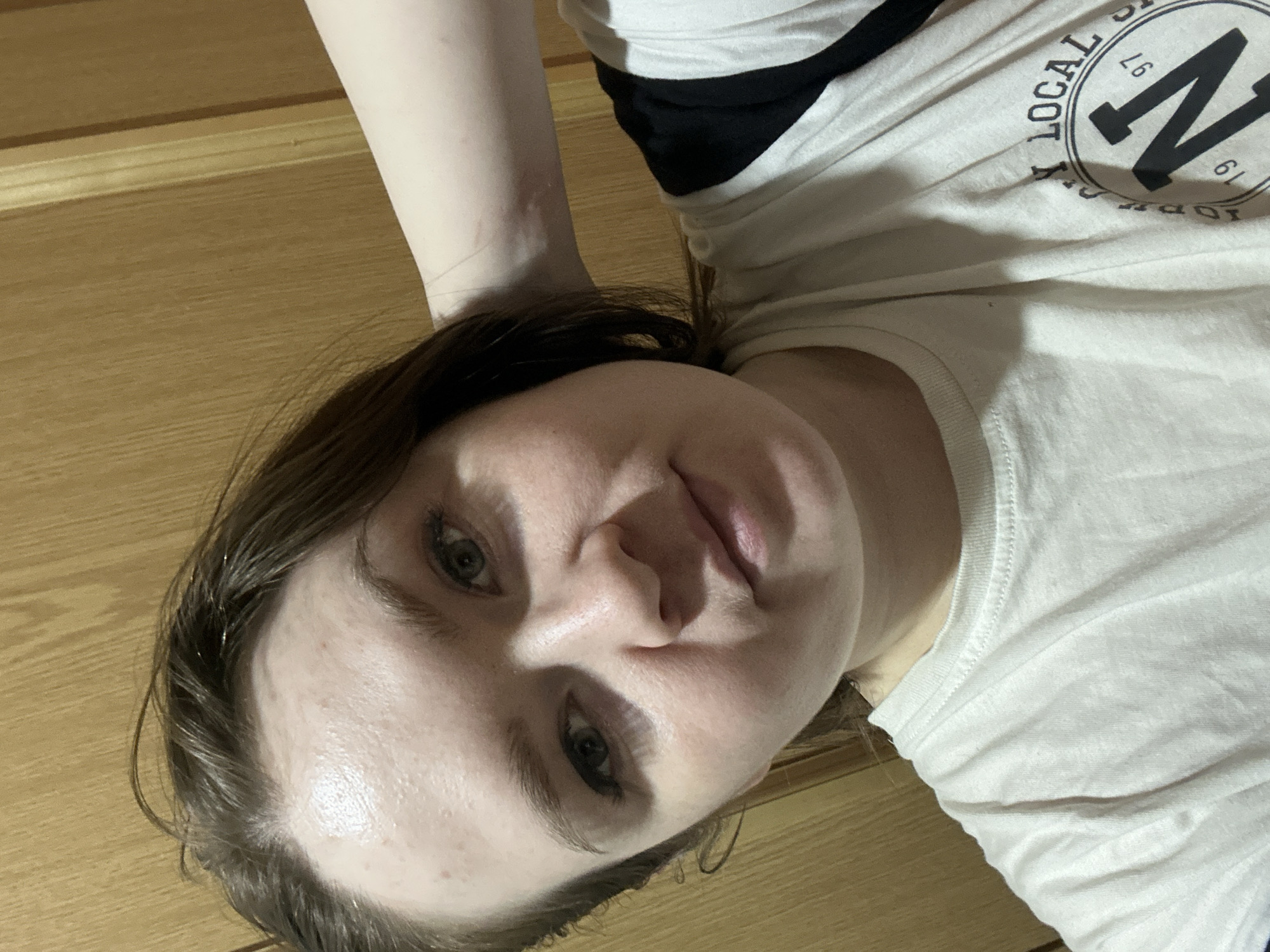 A young woman is lying down, her head tilted slightly towards the camera. Her face is clear, with visible skin texture and details in her eyes, nose, and lips. She is wearing a white t-shirt with a black design that includes the words "LOCAL SUPPLY" and the number "97" and "19". Her arm is visible at the top left of the image. The background consists of a light brown wooden surface. There is no indication of the location being in Chaikovsky, Russia.
