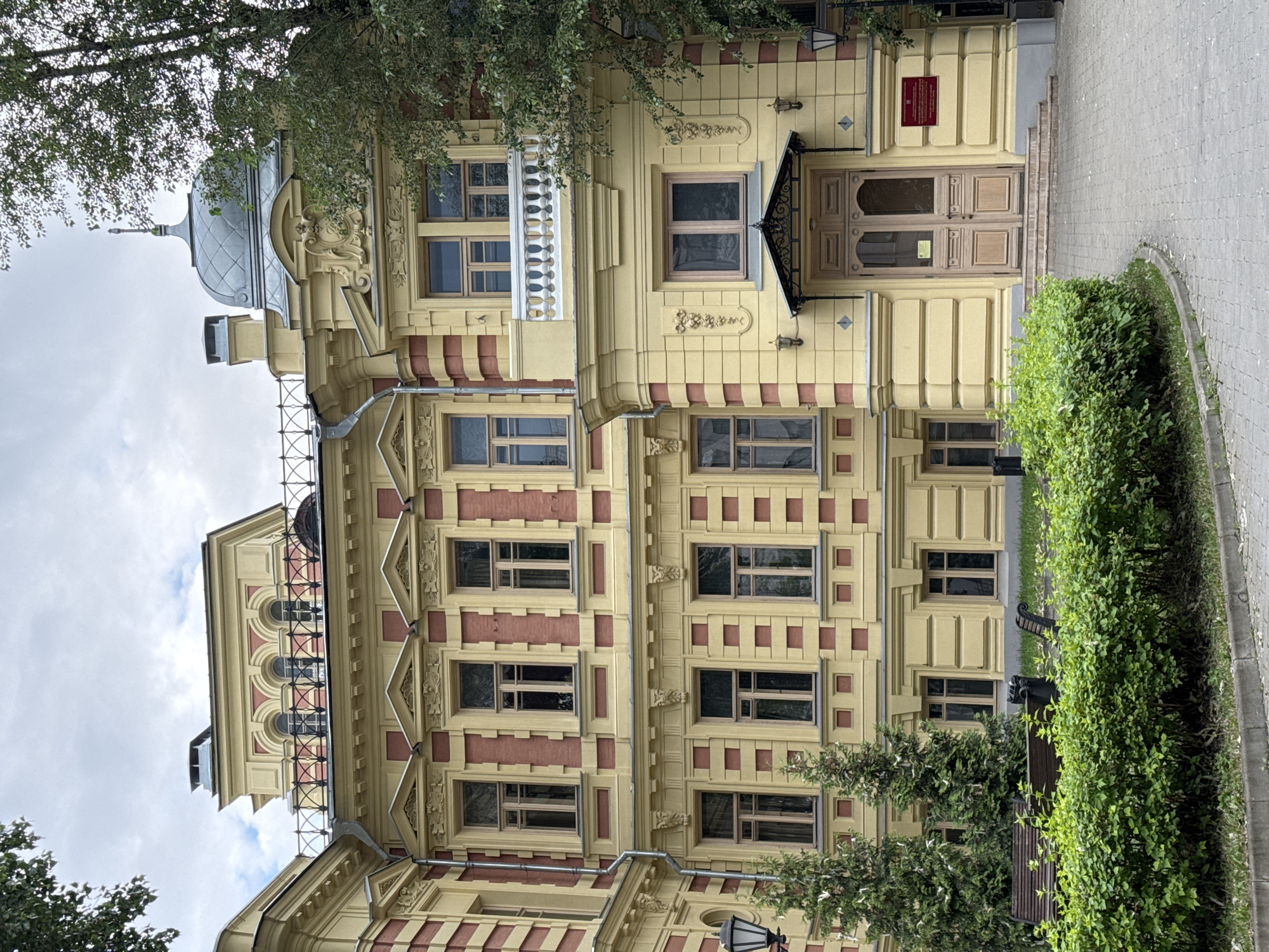 The image shows the facade of a yellow building with red accents, possibly a historical or institutional building, located in Moscow, Russia. The architecture features multiple stories with numerous windows, decorative moldings, and a prominent balcony. A grand entrance with double wooden doors is situated at ground level, approached by a set of stairs. The building is partially surrounded by greenery, including trees and bushes, and there are benches in the foreground. The sky is overcast, suggesting a cloudy day. There are no people or animals visible in the image. The scene appears to be a public space, possibly a park or a courtyard adjacent to the building.