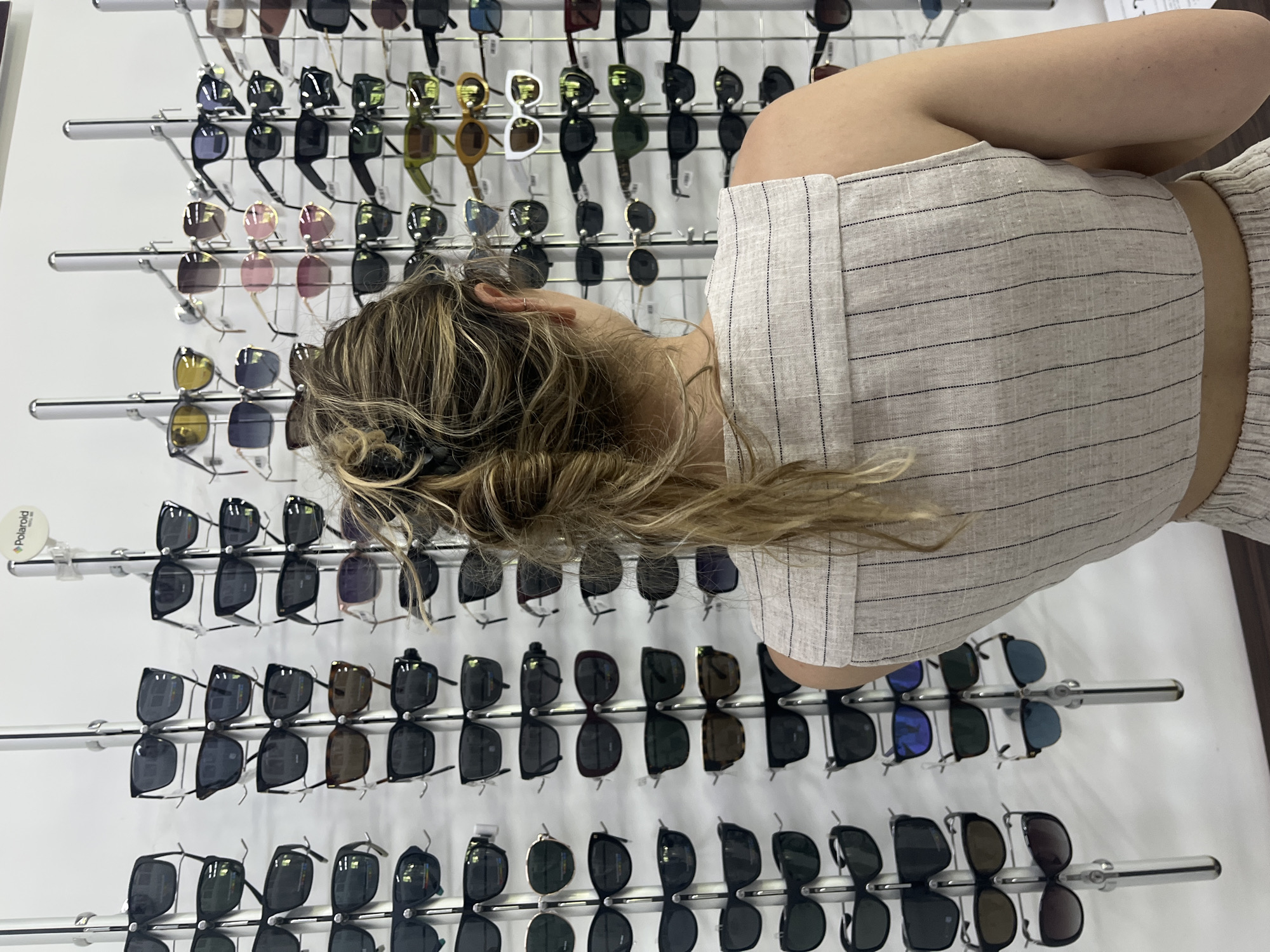 The image presents an overhead view of a person browsing a display of sunglasses within a retail setting in Blagoevgrad, Bulgaria. The subject, seen from behind and above the waist, has light-colored, wavy hair and is wearing a light-toned top with fine dark stripes. Her head is tilted downwards and to the left, indicating focus on the merchandise.
The background consists of a brightly lit, white wall covered with multiple rows of sunglasses displayed on metallic racks. There is a wide variety of sunglasses, showcasing different frame designs, colors, and lens tints, including black, brown, grey, pink, and yellow. The frames appear to be primarily plastic and metal. Many pairs are dark-lensed. The display is well-organized, with reflections of the sunglasses visible on the white shelves beneath each row.
A small, white, circular tag bearing the "Polaroid" brand logo is visible on the upper left side of the display, identifying one of the brands being sold. The scene suggests a typical retail environment, likely an optical shop or a section of a department store dedicated to eyewear, during what appears to be daytime hours due to the bright, even lighting. The person is engaged in the activity of shopping for or inspecting sunglasses.