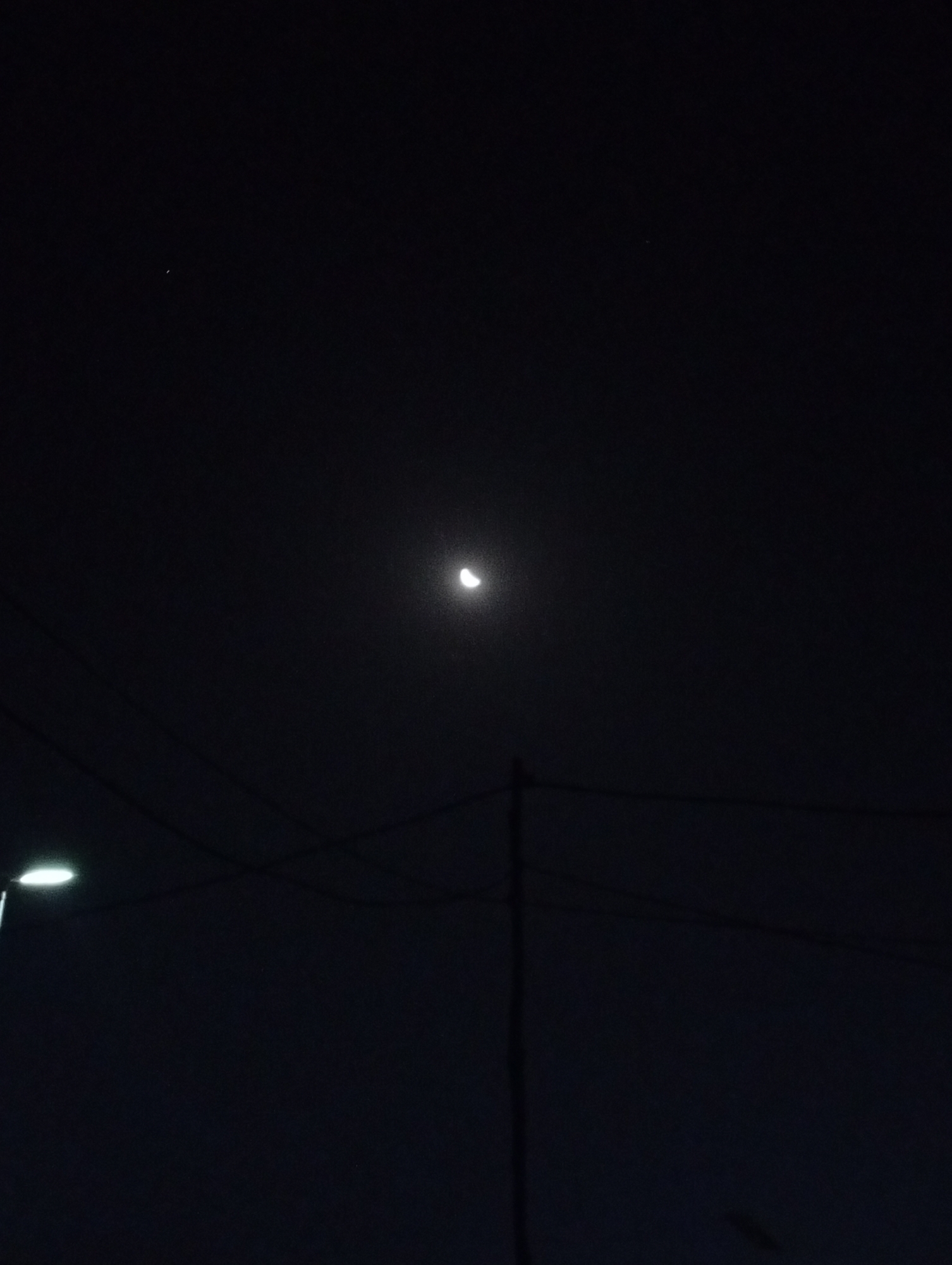 A night scene in Adankolo, Nigeria. A crescent moon is centered in the upper portion of the frame. A small, faint star is in the upper left corner. Below, a street light illuminates the lower left. Power lines and a utility pole are visible in the foreground. The overall scene is dark.
