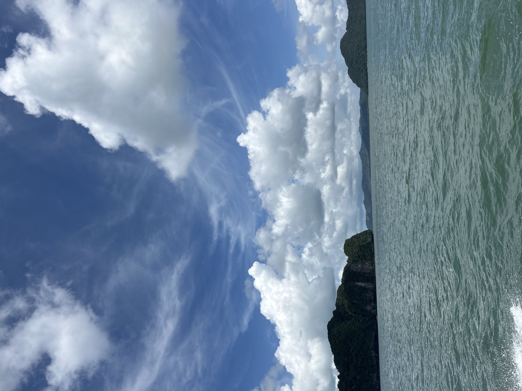 The image depicts a body of water with a view of lush green islands in the distance. The water is a deep blue-green, with visible ripples and a small amount of white foam in the lower left corner, suggesting movement, possibly from a boat. Above the water, the sky is a vibrant blue with a mix of fluffy white cumulus clouds and thinner, wispy cirrus clouds. The islands are covered in dense vegetation and appear to be mountainous. The overall scene conveys a tropical or coastal environment, characteristic of locations like Krasom, Thailand, known for its beautiful islands and seascapes. No people or specific structures are visible.