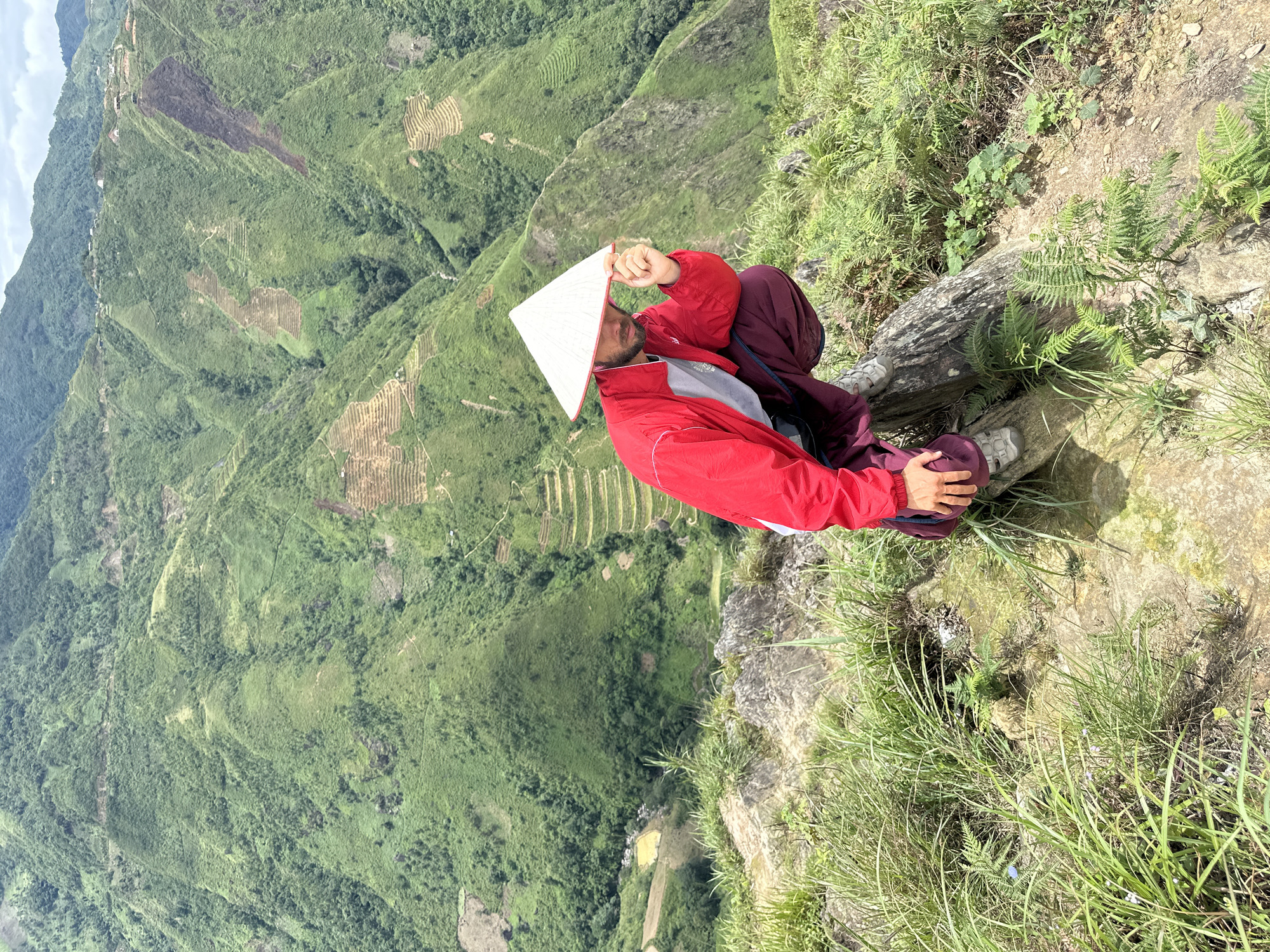 A man wearing a red jacket, burgundy pants, hiking boots, and a conical hat squats on a rocky, grassy outcrop overlooking a lush, terraced mountain landscape in Ta Xua, Vietnam. The background reveals steep, verdant mountains with visible agricultural terraces and a winding path. The man is holding the brim of his hat with his right hand, and his gaze is directed downwards. The foreground consists of rocks, green grass, and ferns. The sky is overcast.