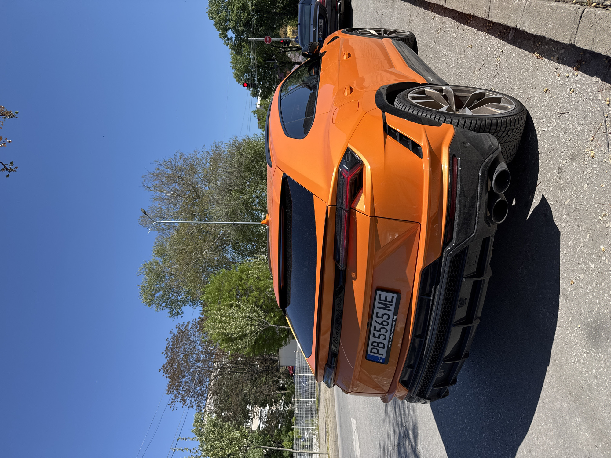 An orange Lamborghini Urus is parked on a street in Plovdiv, Bulgaria. The rear of the vehicle is visible, including the license plate "PB 5565 ME." The sky is blue, and there are trees in the background. Another vehicle is partially visible in the background, as well as a street lamp and traffic light. The street is paved and has some debris.