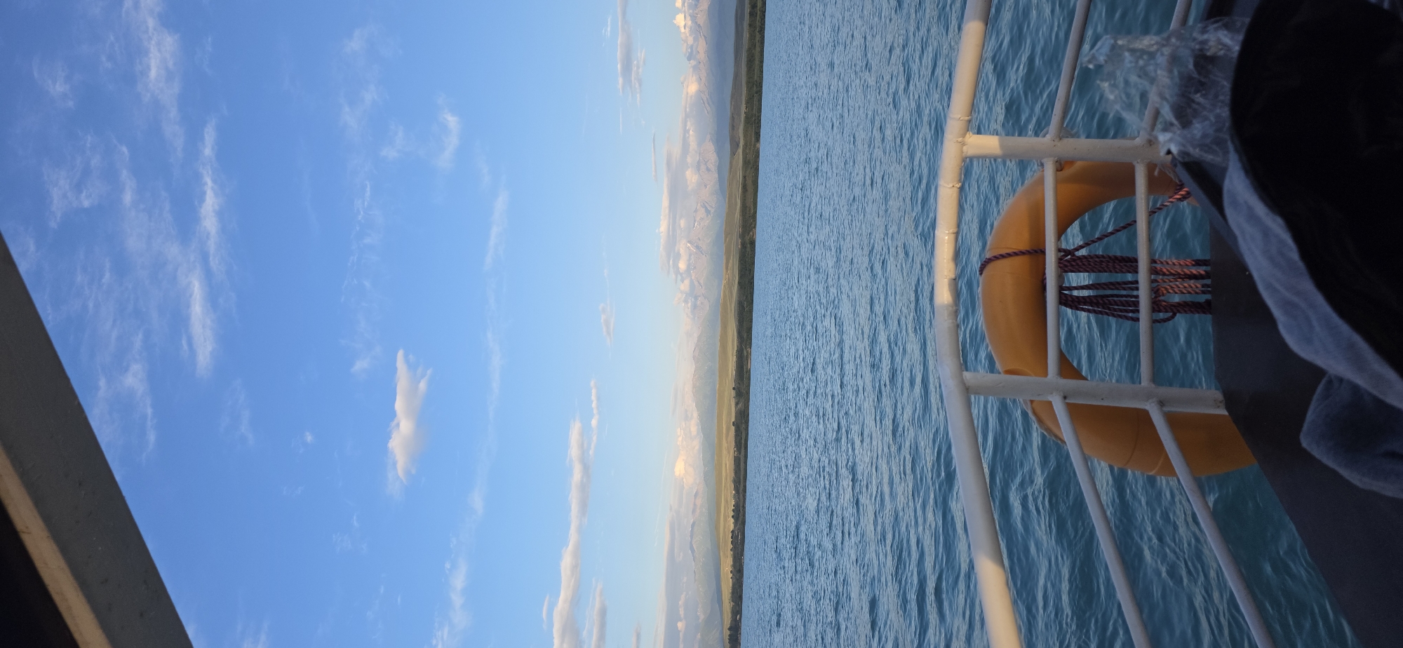 This media file captures a scenic view from a boat on a body of water. The sky is a vibrant blue with scattered wispy white clouds, suggesting a clear day. In the distance, a long stretch of land is visible, characterized by rolling hills and mountains that appear to be illuminated by the soft light of either sunrise or sunset. The water is a deep blue, with gentle ripples indicating calm conditions.
On the right side of the frame, a portion of the boat is visible, featuring a white metal railing. Attached to this railing is a bright orange life preserver, secured with a purple and red rope. A dark object, possibly a bag or clothing, is draped over the railing near the life preserver. The scene conveys a sense of tranquility and natural beauty. The time of day appears to be either early morning or late afternoon due to the soft lighting on the distant mountains. The location is identified as Pristan'-Przheval'sk, Kyrgyzstan, which is a port city on the eastern shore of Lake Issyk-Kul. There is no visible text in the image.