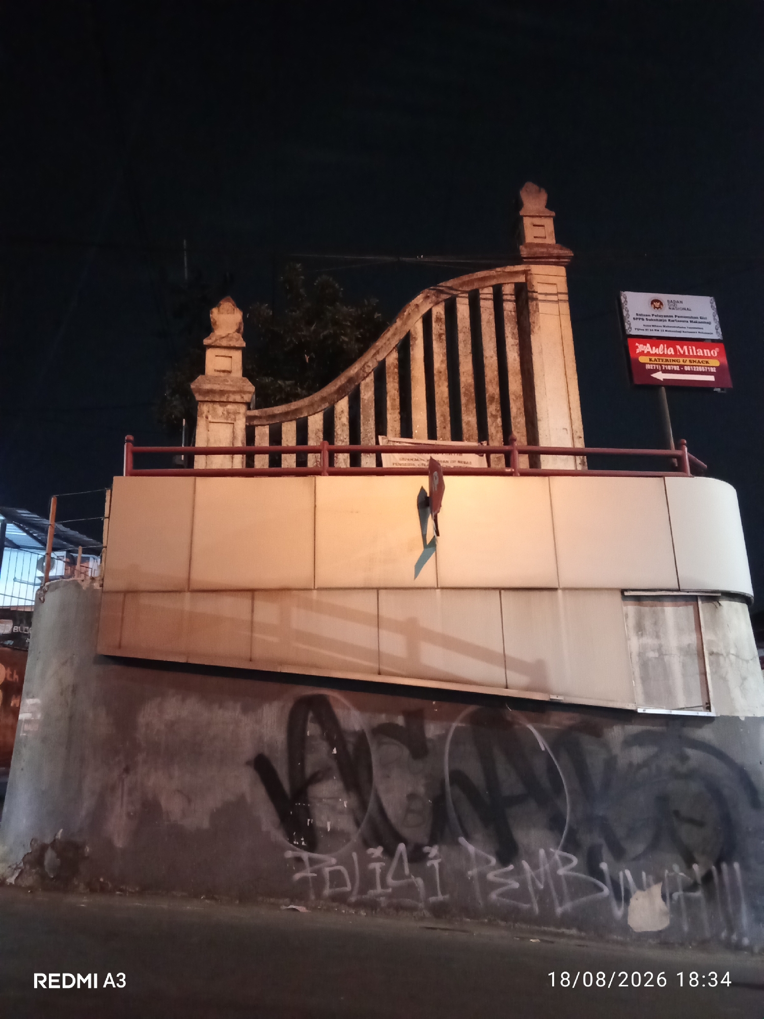 The image depicts a night scene in Purbayan, Indonesia. A concrete structure, possibly part of a building or monument, is illuminated by artificial light. The structure features a decorative stone balustrade at the top, with vertical stone pillars and a curved top railing. Below the balustrade, the structure is clad in beige rectangular panels. The lower portion of the structure is a rough concrete wall covered in graffiti. A street sign with text and a red arrow is mounted on the right side of the structure. The environment is dark, suggesting it is nighttime, with visible overhead electrical wires. The bottom left corner of the image displays the text "REDMI A3", and the bottom right corner shows the date "18/08/2026" and time "18:34".